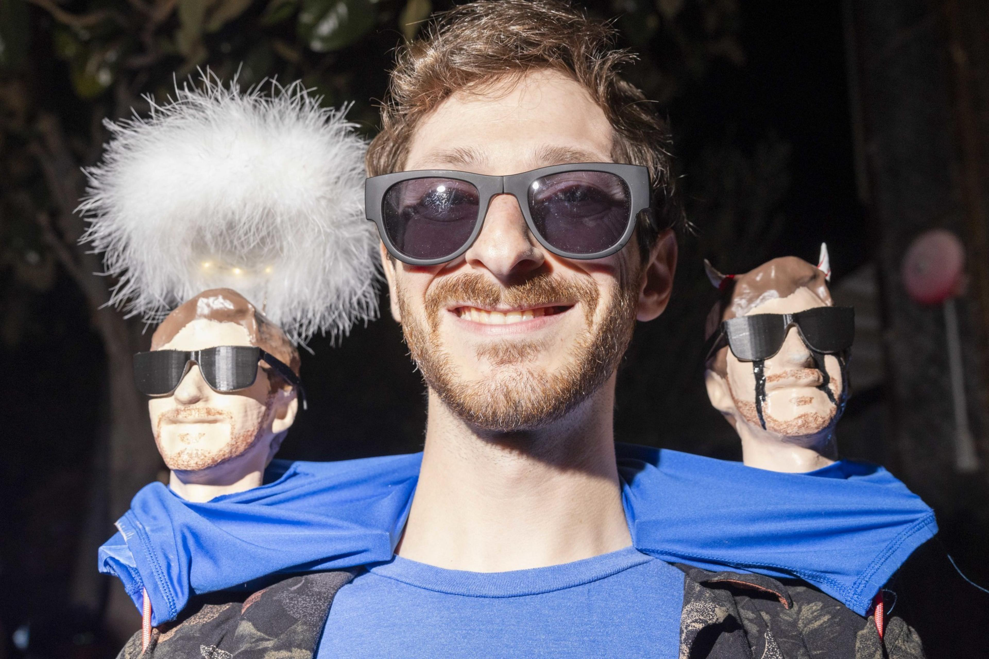 A man wearing sunglasses smiles, flanked by two small models of himself. One model has angelic features, with a fluffy halo, and the other has devil horns.