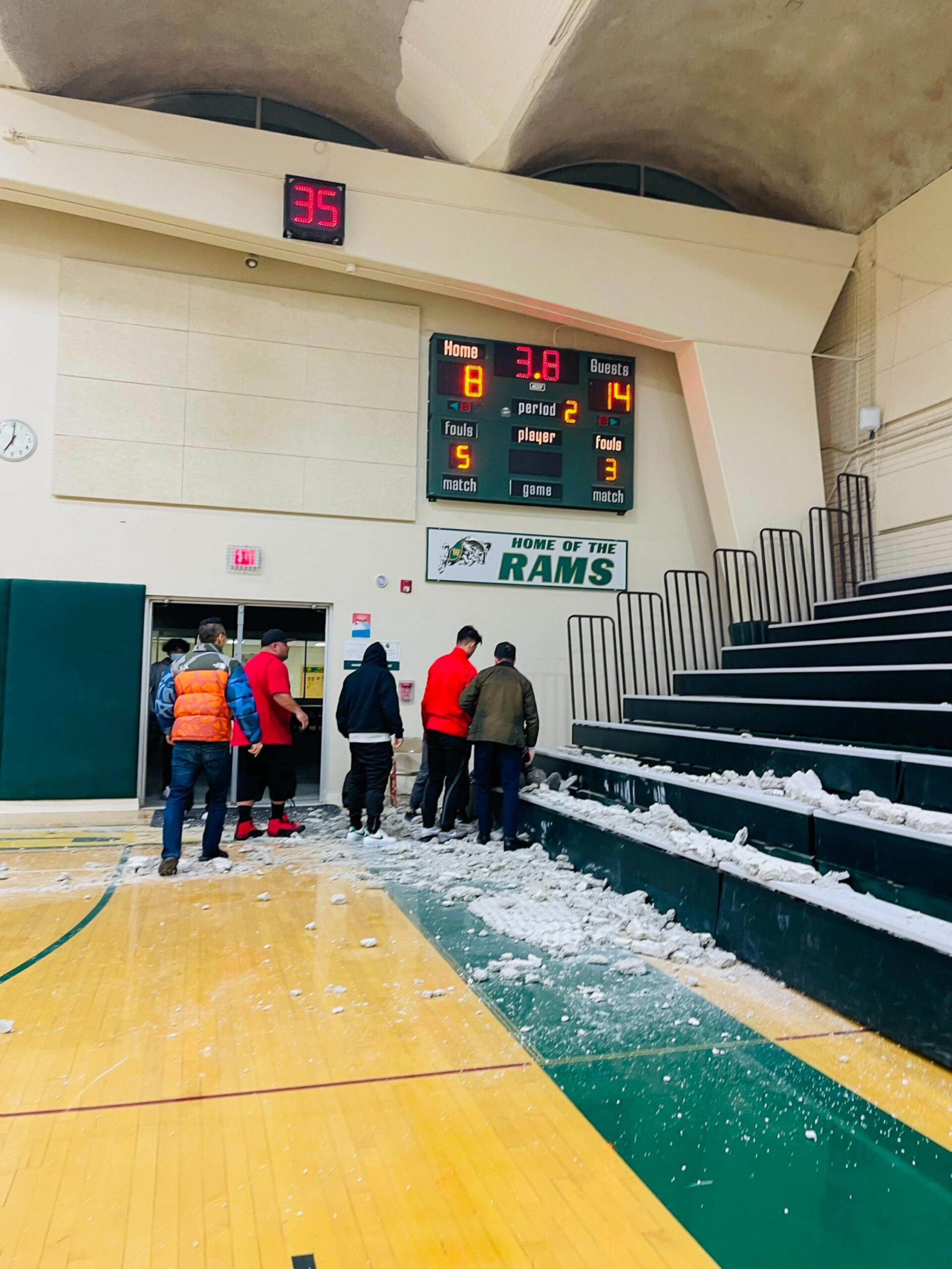 A basketball gym with a scoreboard showing 3.8 seconds left and a guest lead of 14-8, with several people inspecting debris scattered on the bleachers and floor.