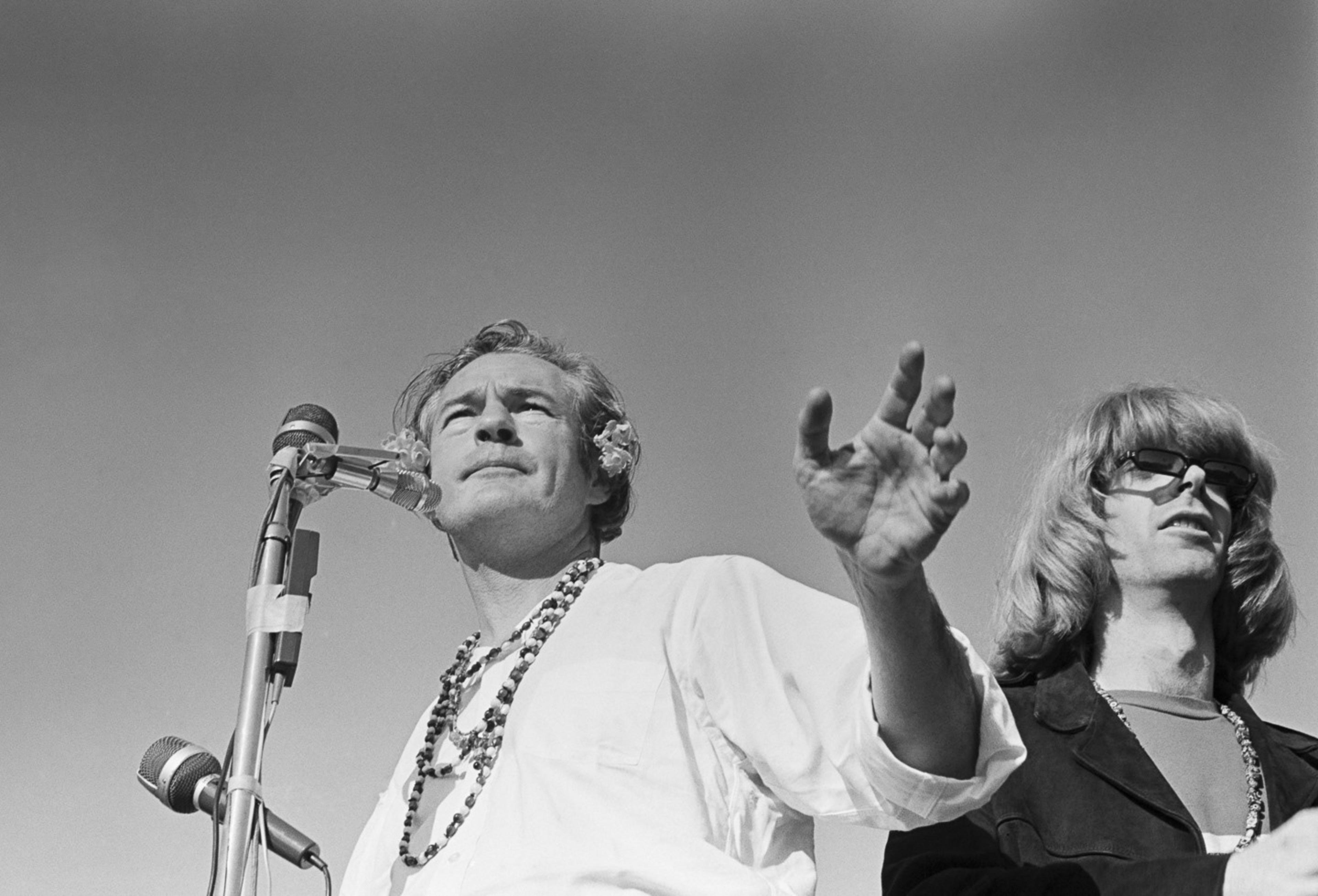 Two men stand at microphones. The man on the left is speaking passionately with flowers in his hair and wearing a necklace. The man on the right has long hair and glasses.
