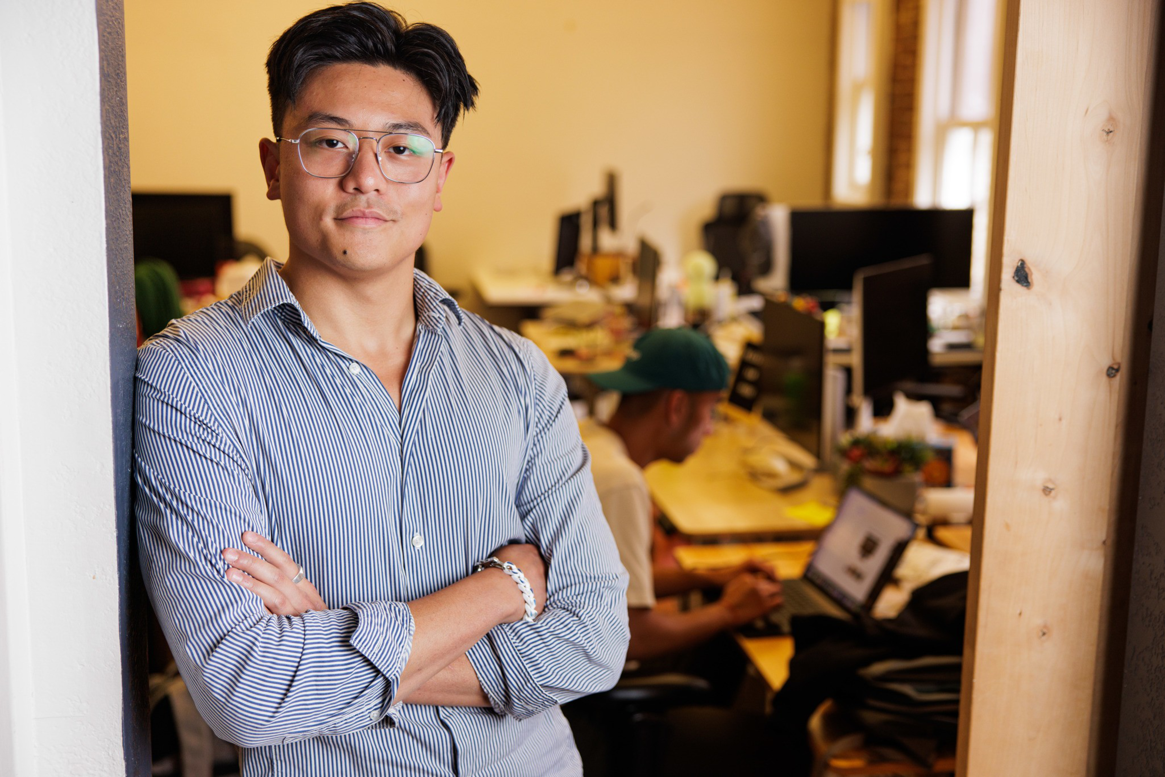 A man wearing glasses and a striped shirt stands with arms crossed in an office while another person works on a laptop in the background.