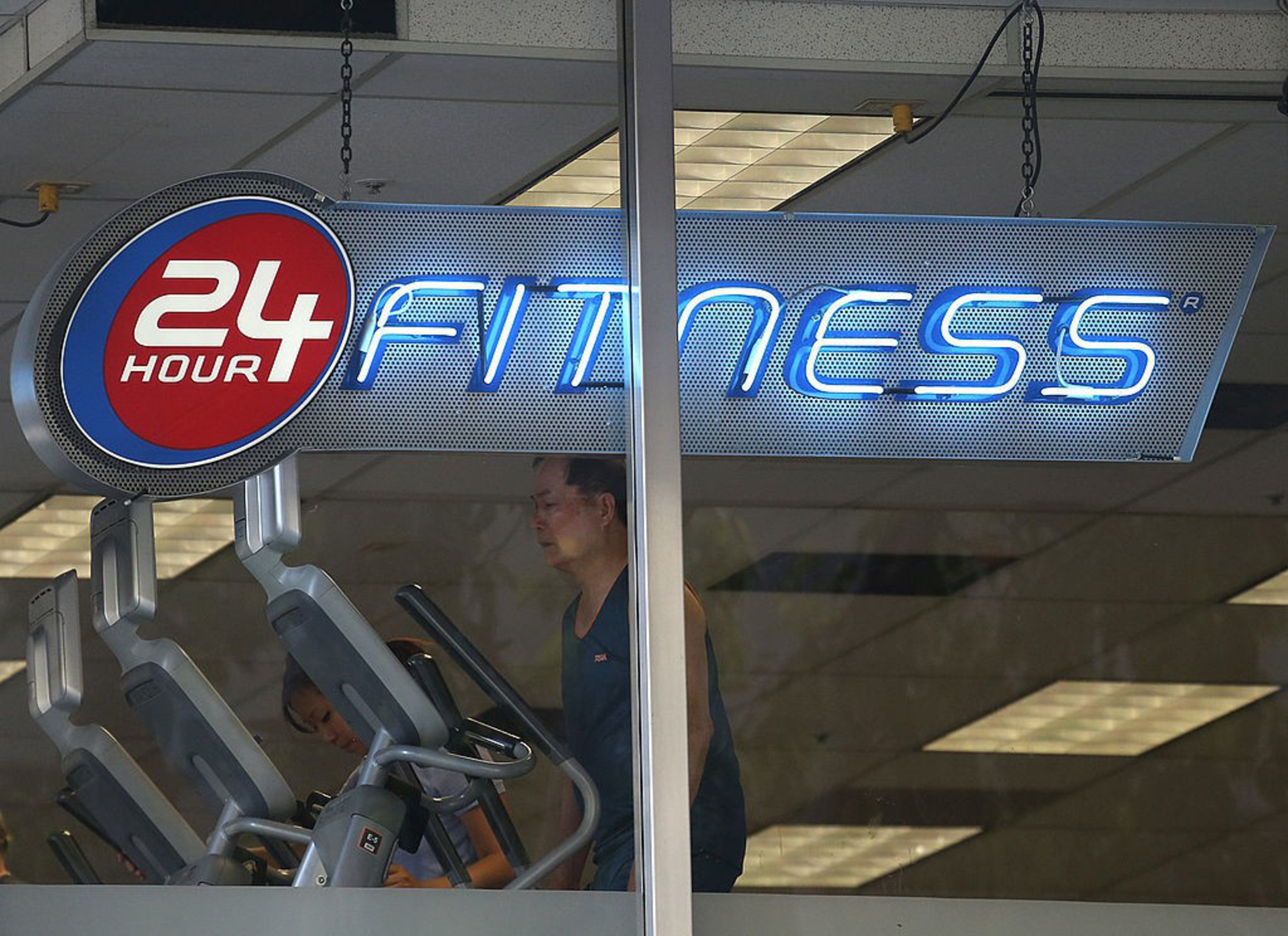 A man exercises on a treadmill inside a 24 Hour Fitness gym, visible through a glass window beneath a bright neon gym sign.