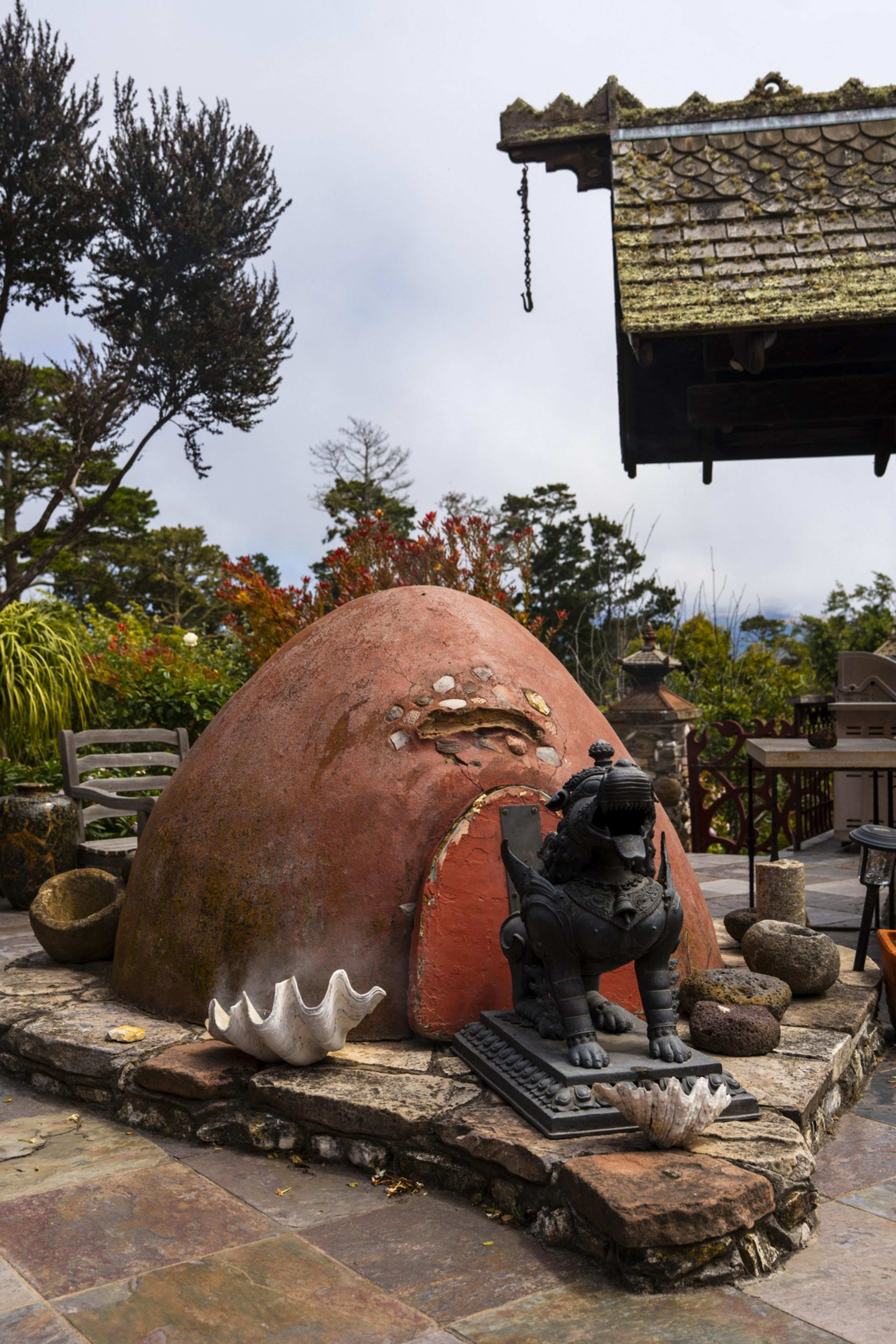 A rustic outdoor setting features a dome-shaped stone structure, a black lion statue, and large seashells. Lush greenery and an ornate roof decorate the background.