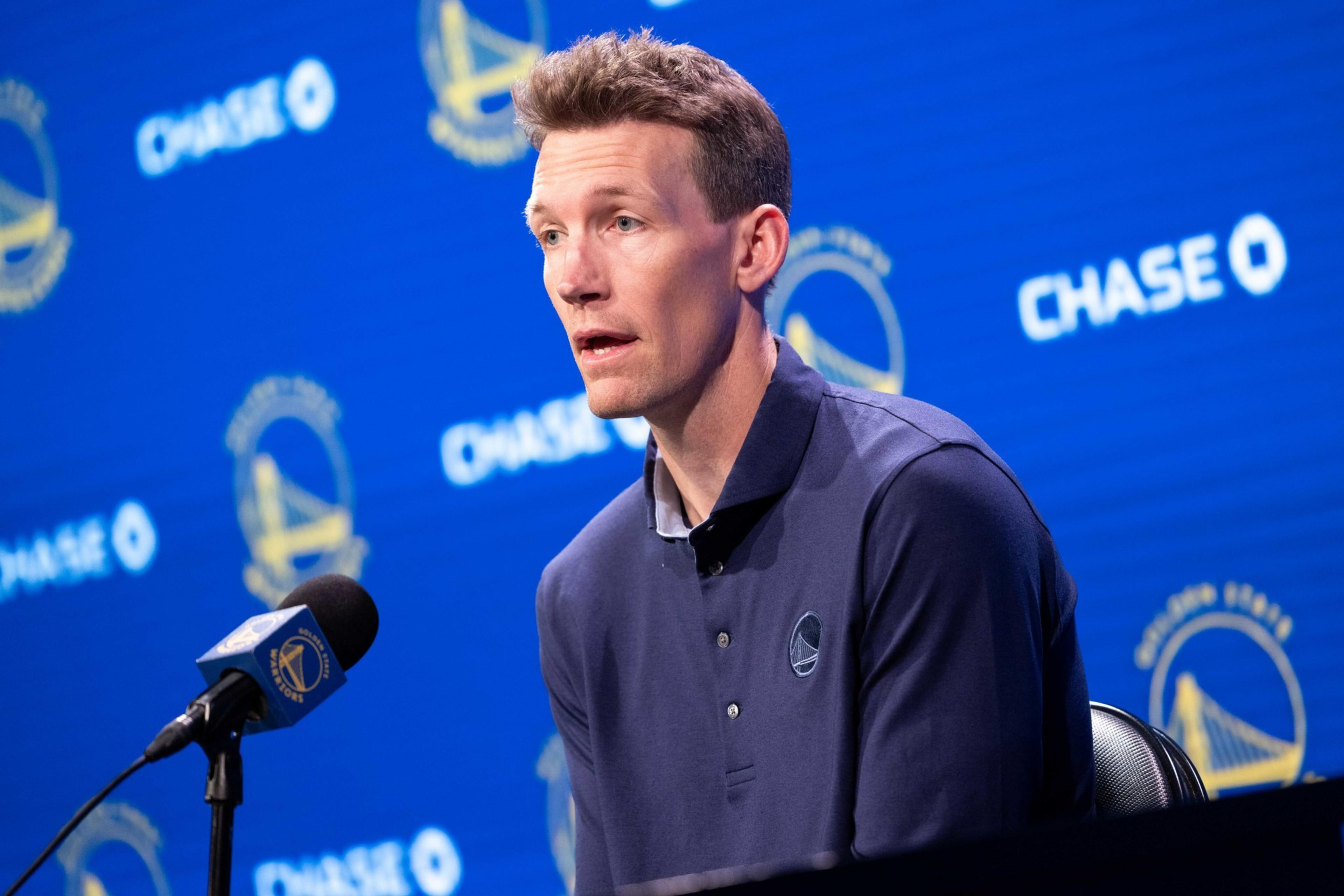 A man in a navy shirt sits at a press conference with a microphone. The background features Golden State Warriors and Chase logos.