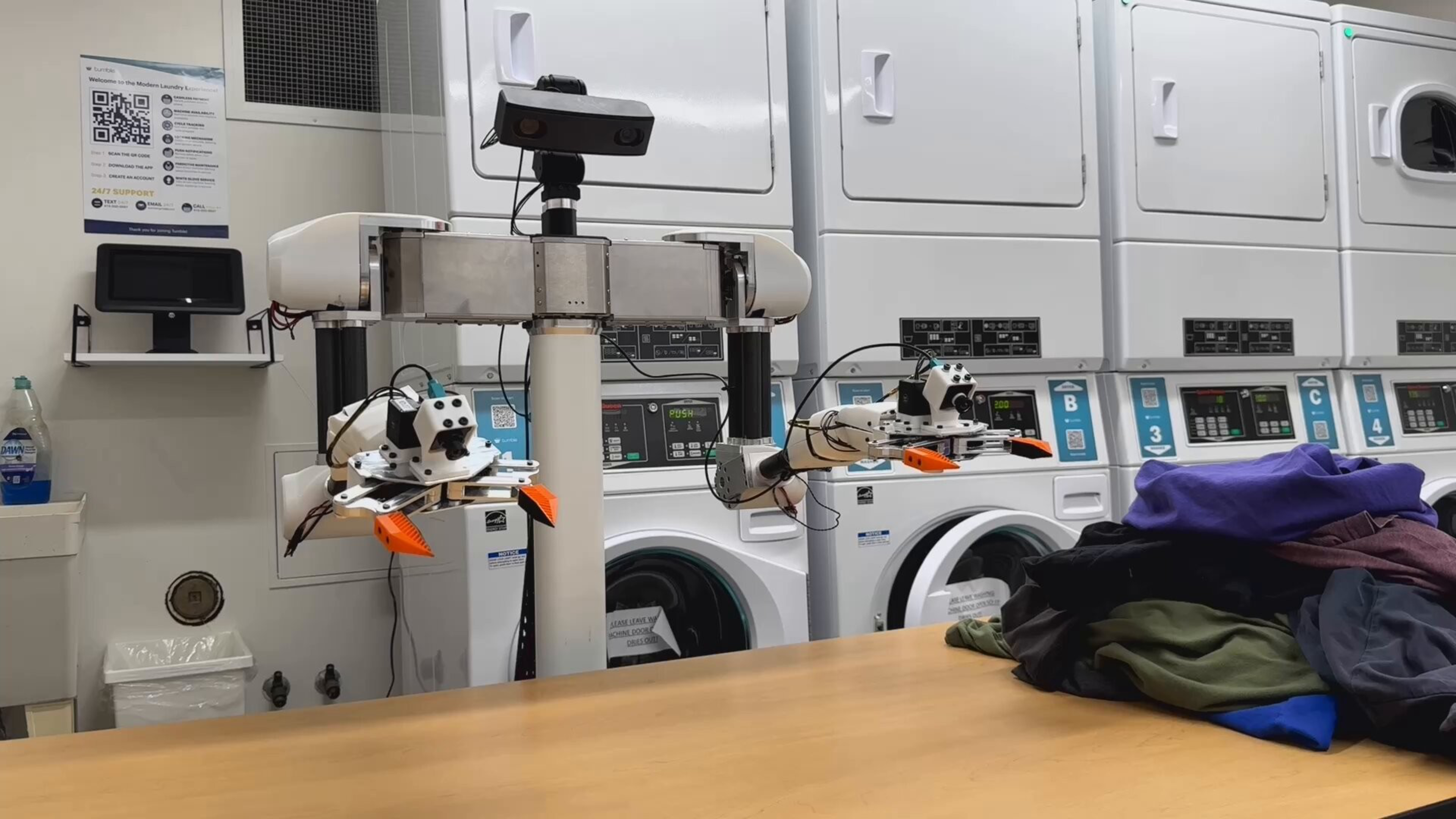 A robot with two claw-like arms stands in front of stacked laundry machines, with a pile of clothes on a wooden table nearby.
