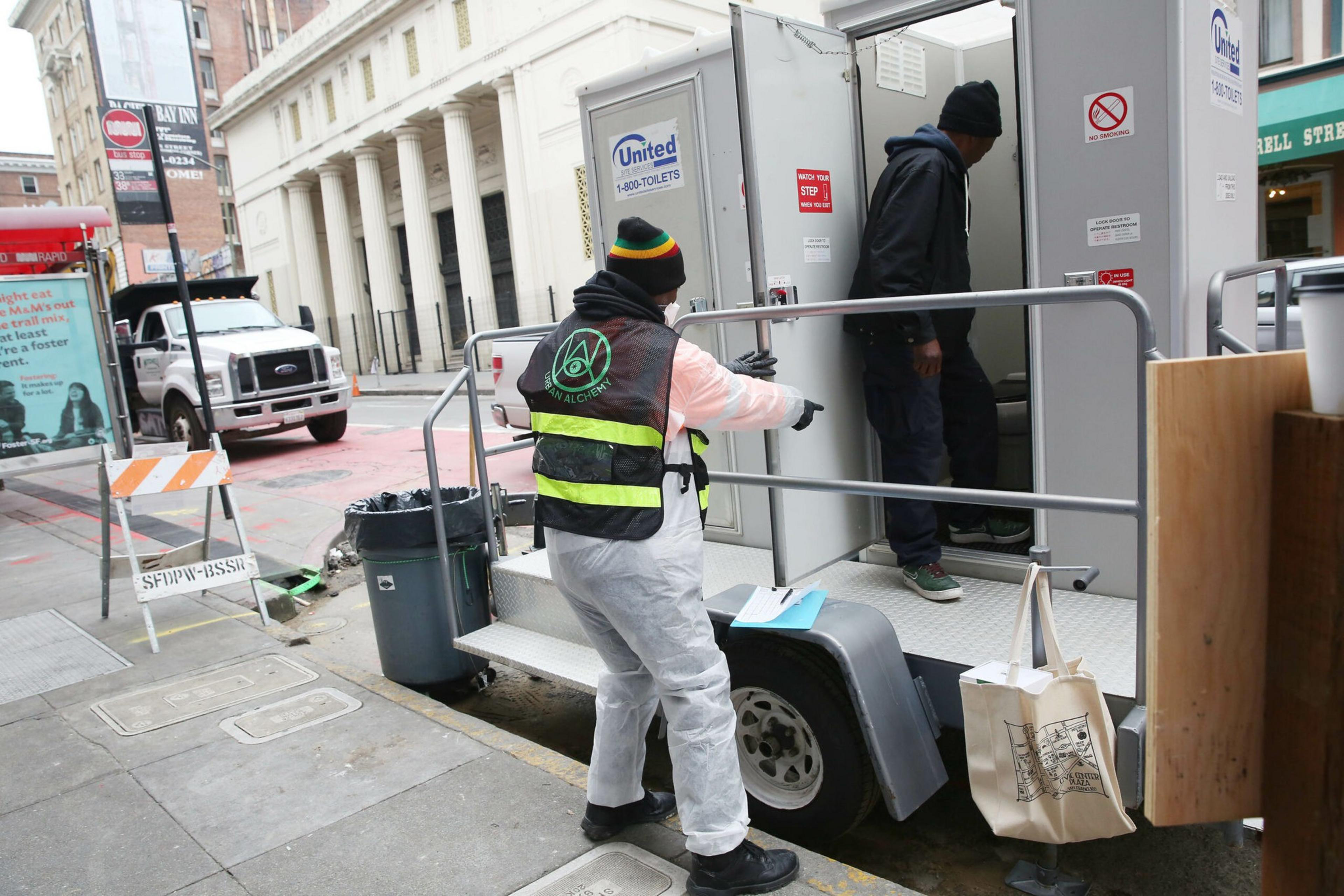 A worker in protective gear guides a man entering a portable restroom trailer on a city sidewalk near buildings and a parked truck.