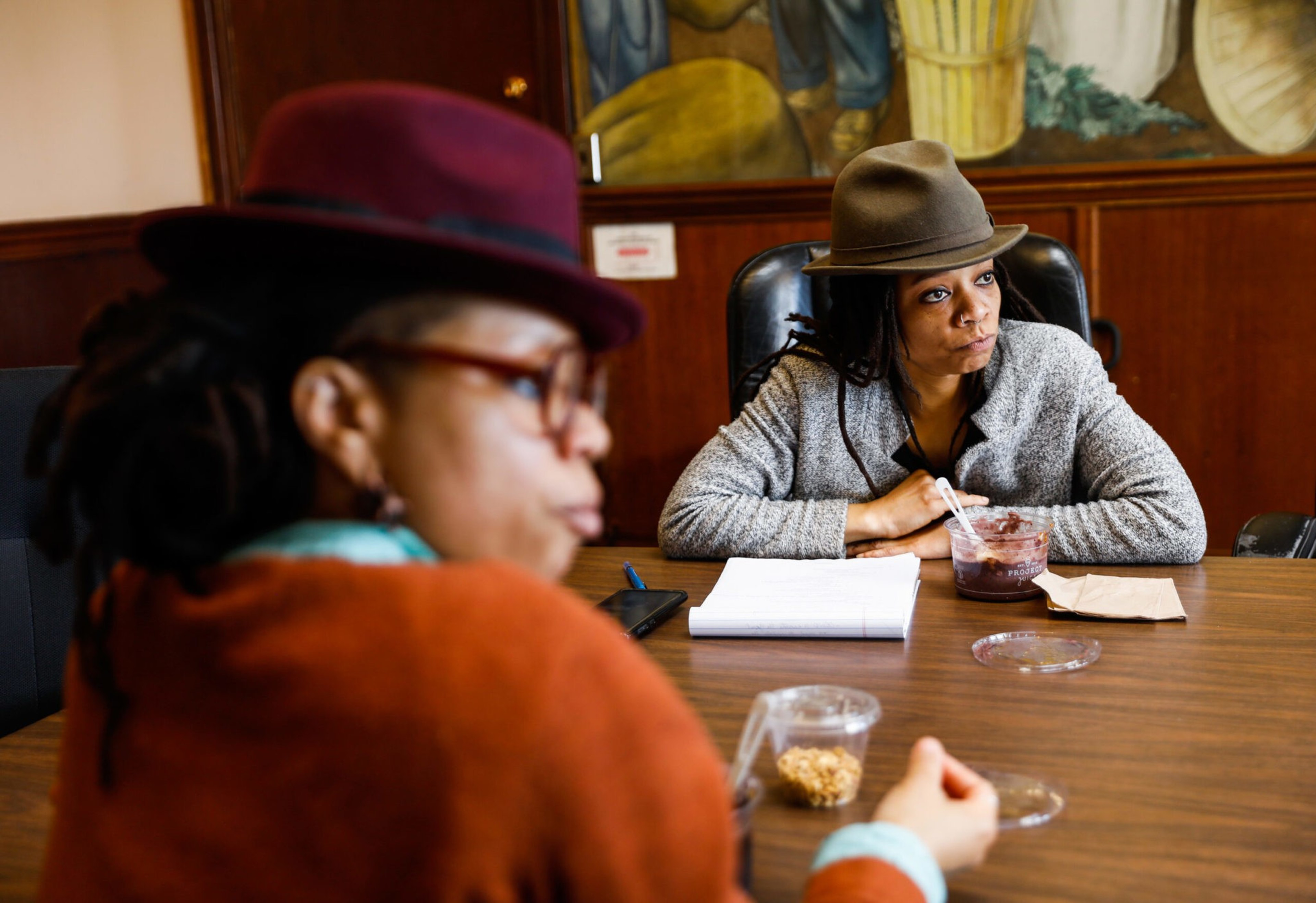 Two women wearing hats sit at a table; one faces forward with a notebook and snack, while the other is blurred in the foreground.