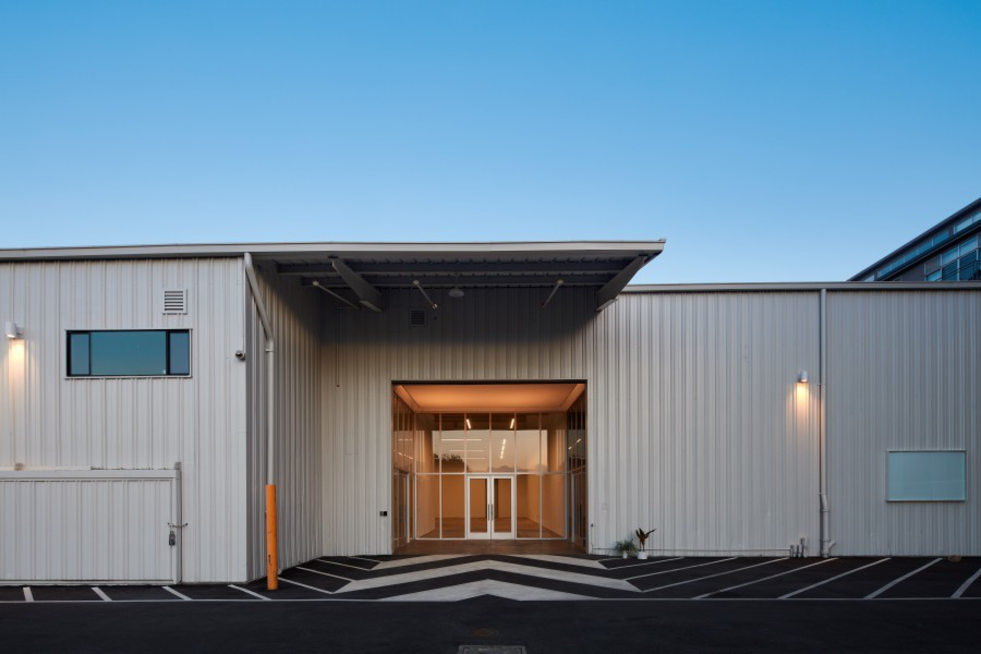 A modern industrial building with a large glass entrance, metal siding, marked parking spaces, and a clear blue sky above.