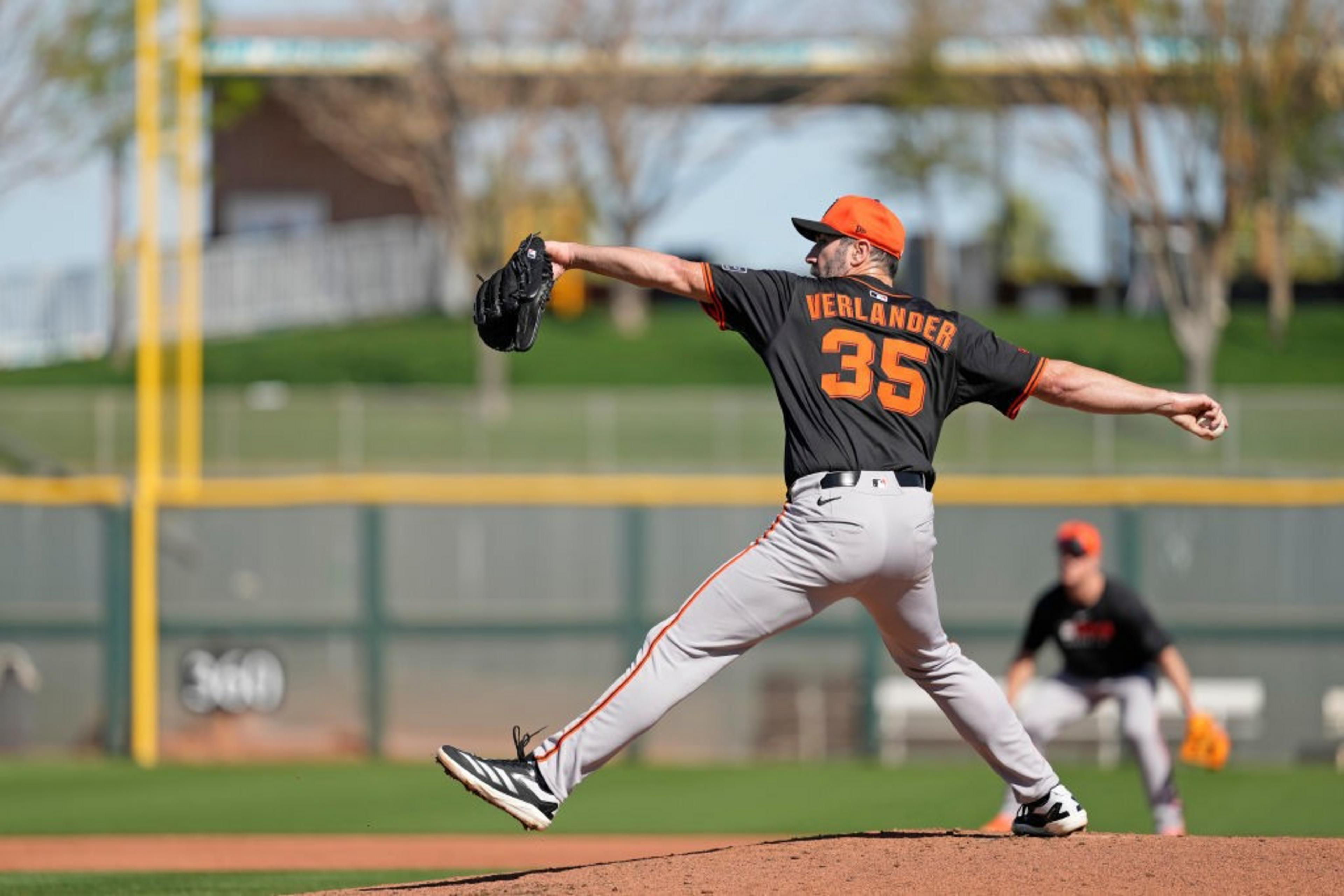 A baseball pitcher in a black and orange uniform is mid-throw on a mound, with a fielder crouching in the background on a sunny day.