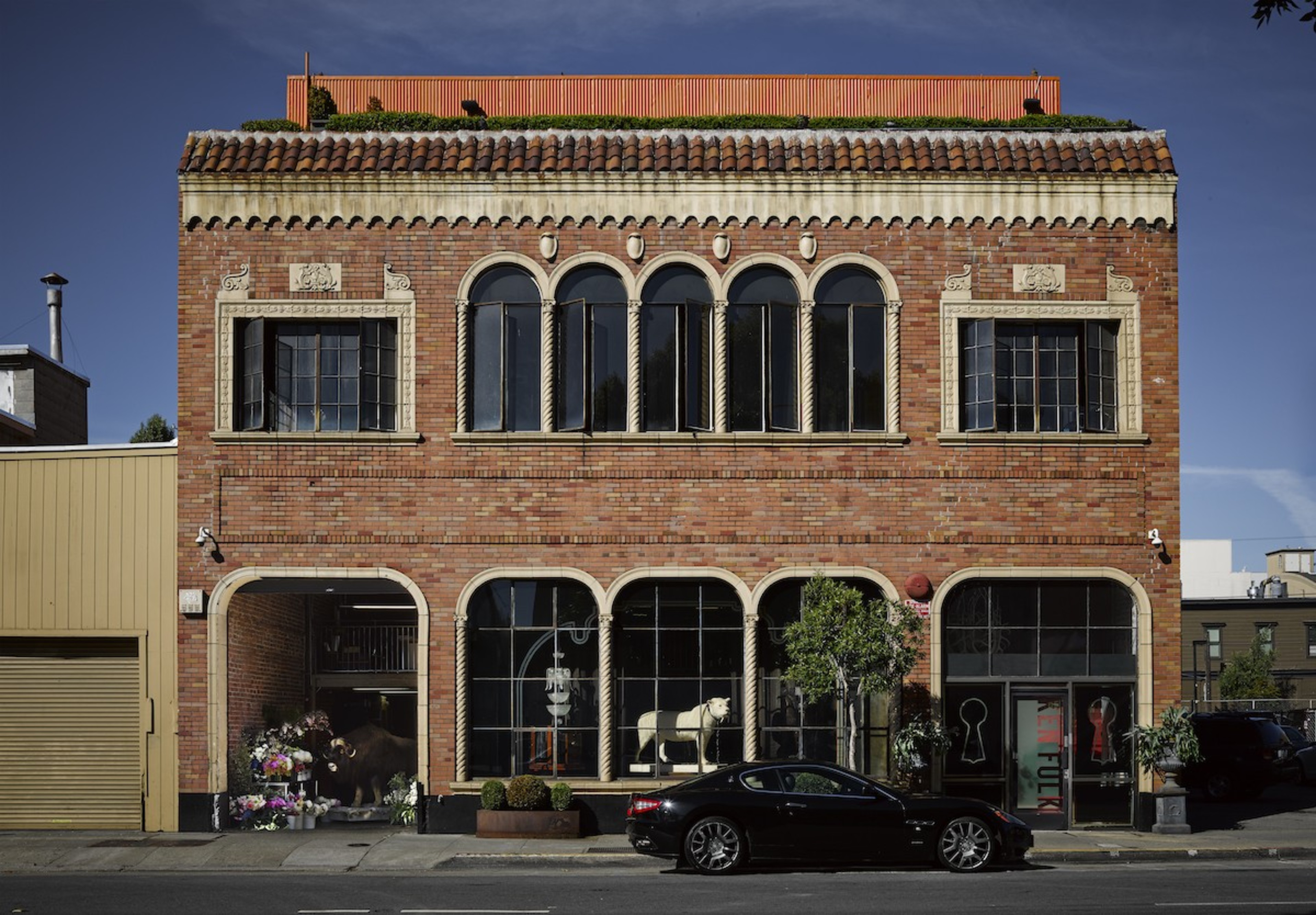 A two-story brick building with tall arched windows and ornate stone trim. The ground floor has a garage with flowers and animal statues, and a black car parked outside.