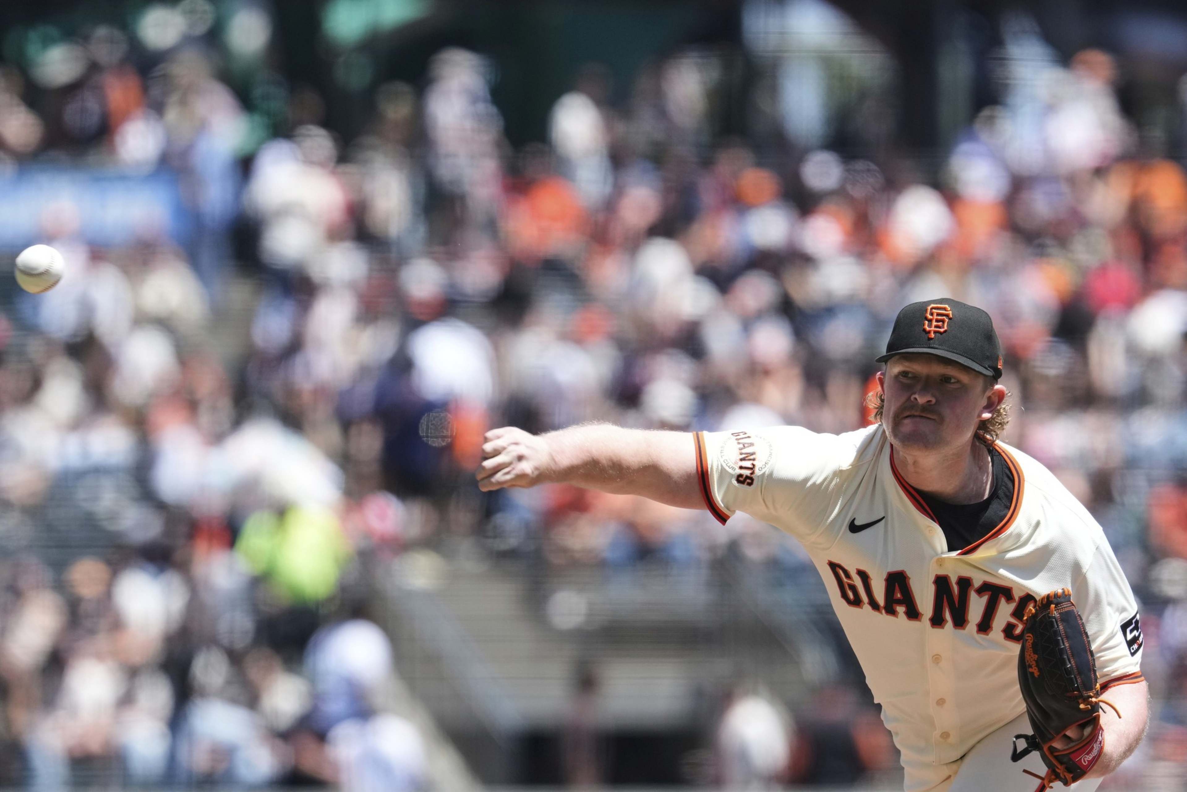 A baseball player in a Giants uniform is pitching on a field. The ball is mid-air, and there's a crowd with blurred figures in the background.