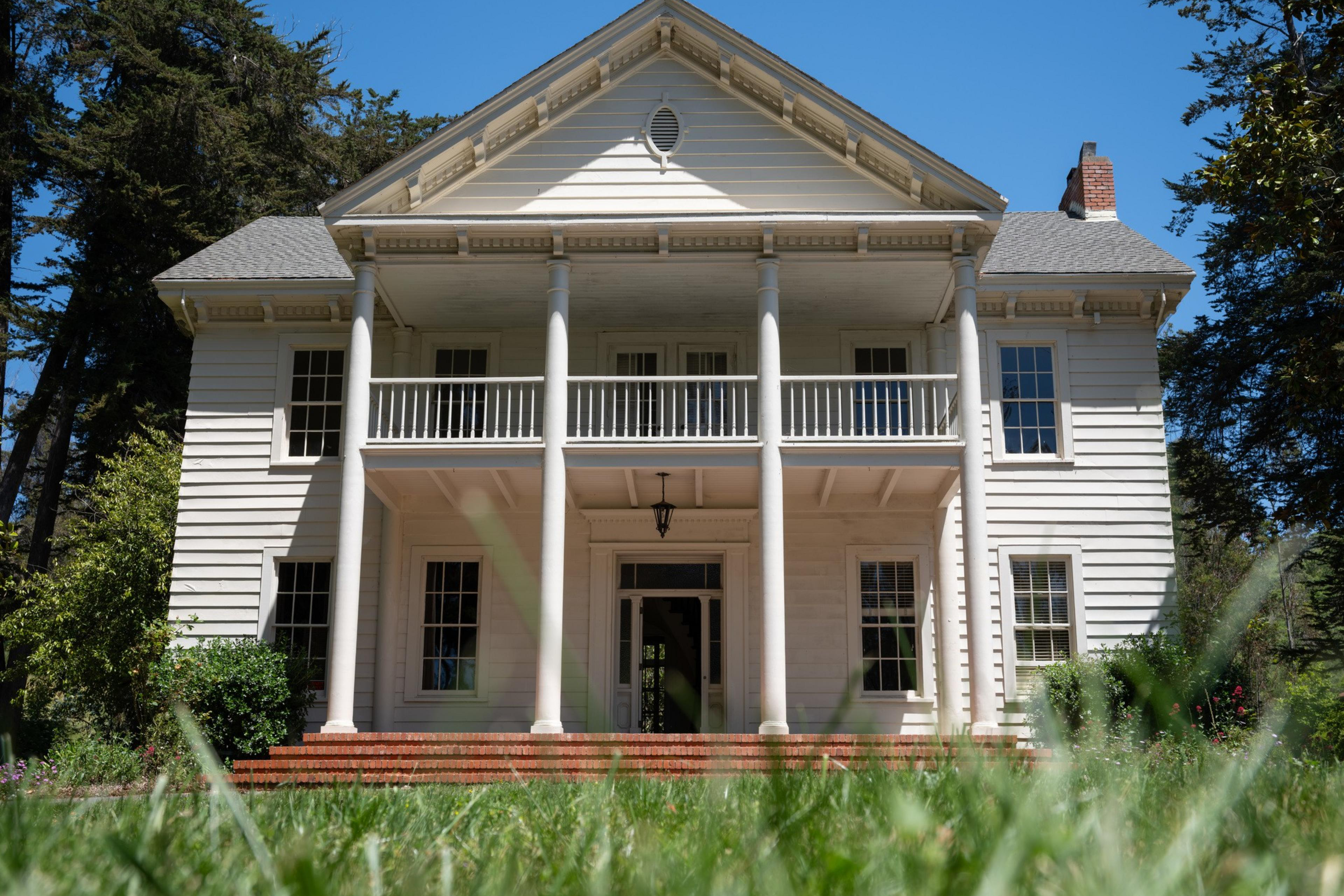 A two-story white house with tall pillars stands against a clear blue sky, surrounded by green trees and shrubs, with a brick step entrance in the foreground.