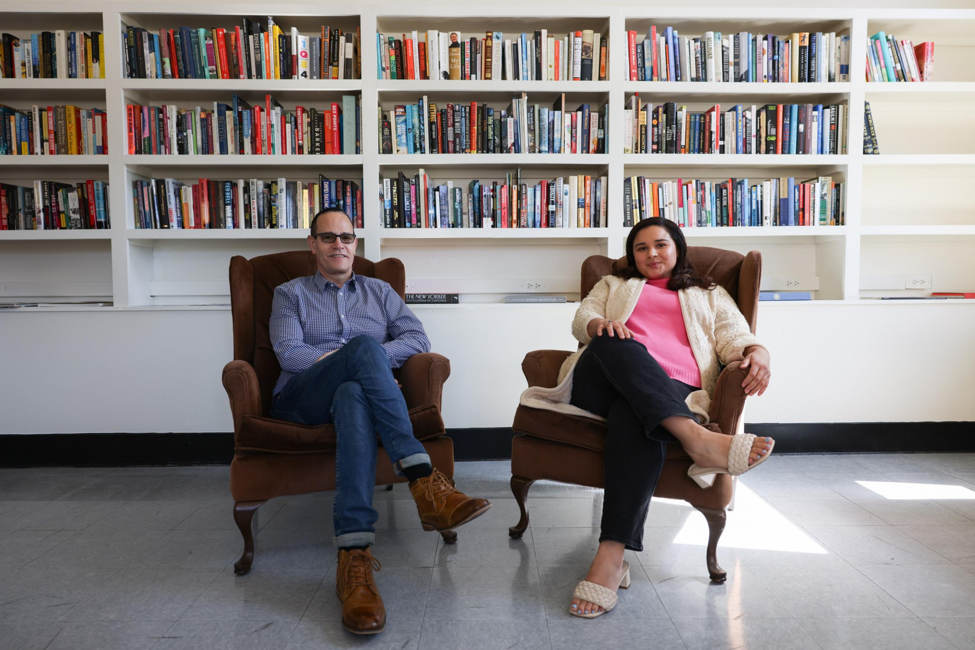 A man and a woman sit relaxed in brown armchairs with crossed legs in front of white bookshelves filled with books.