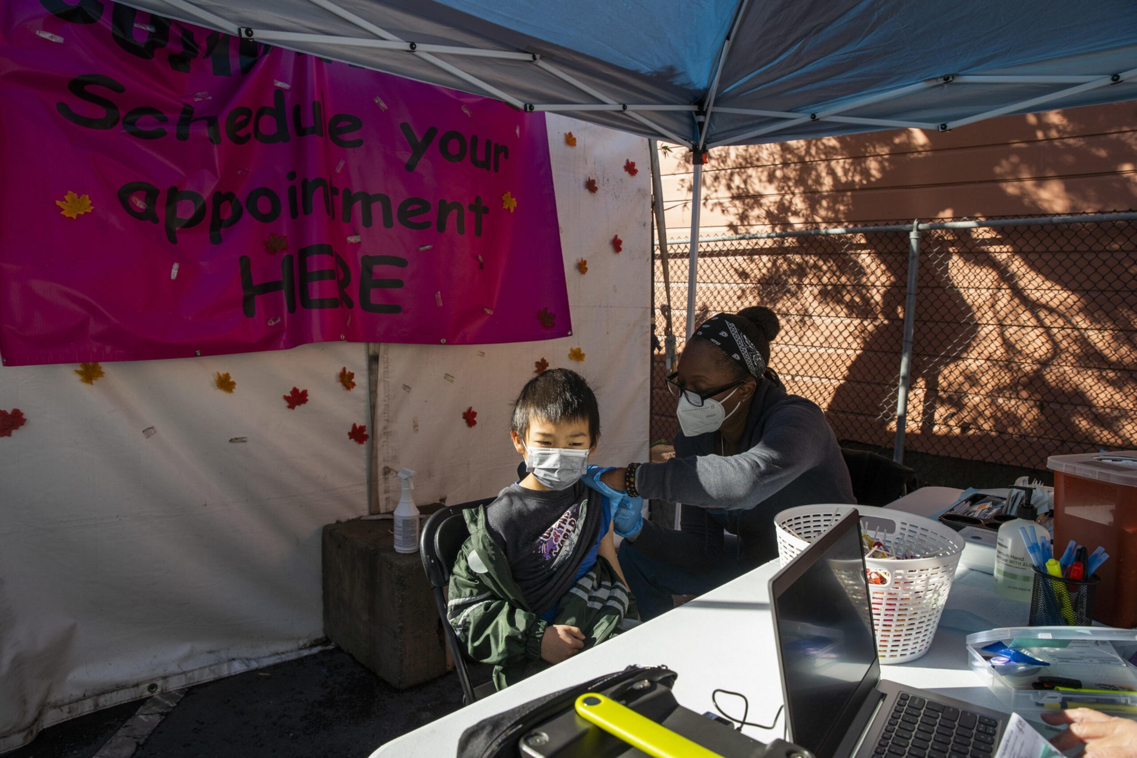 A masked healthcare worker gives a vaccine shot to a masked child seated under a tent with a pink "Schedule your appointment HERE" sign.