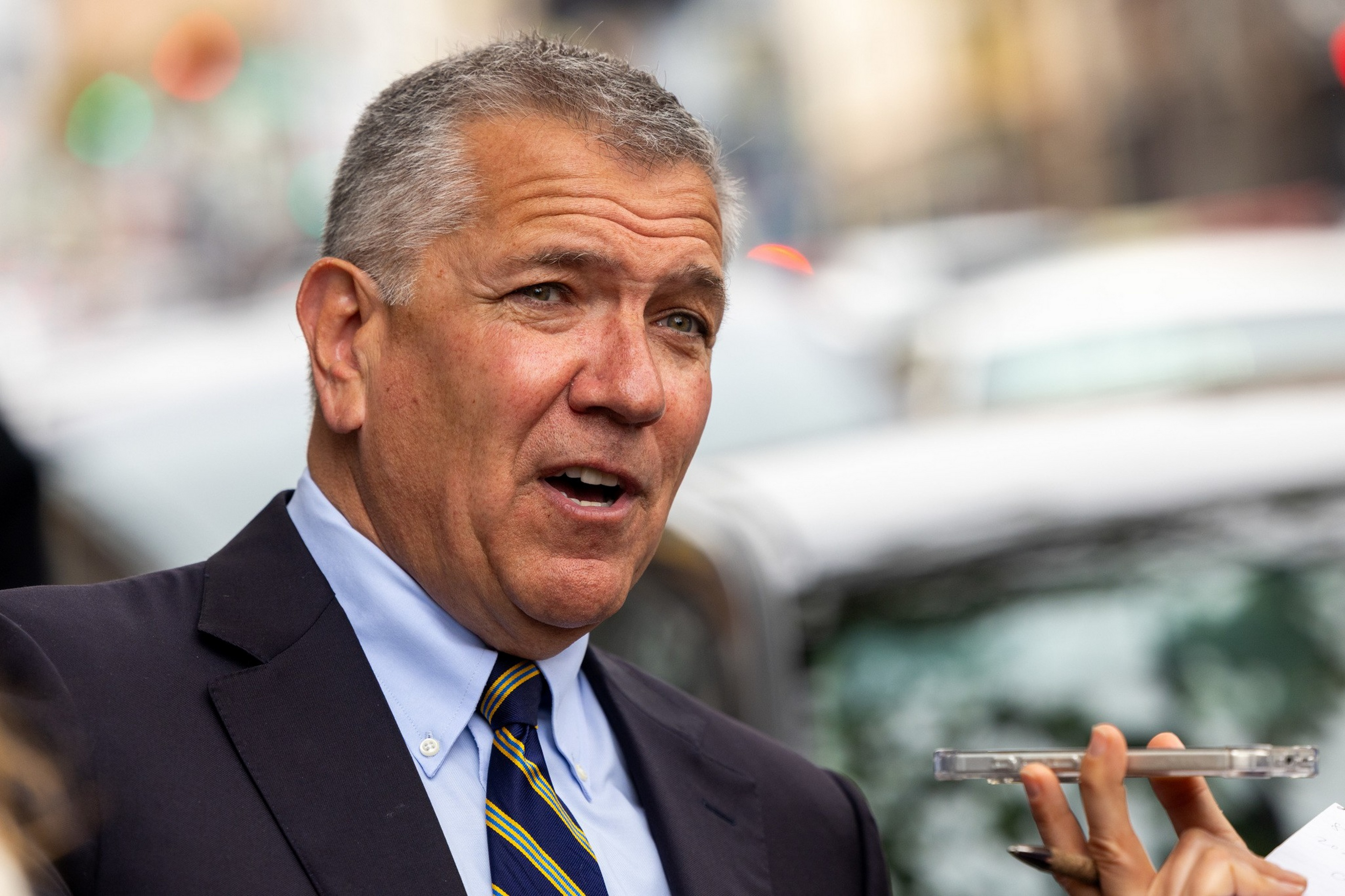 A middle-aged man in a suit and striped tie speaks while a hand holds a smartphone near his face, with blurred background.