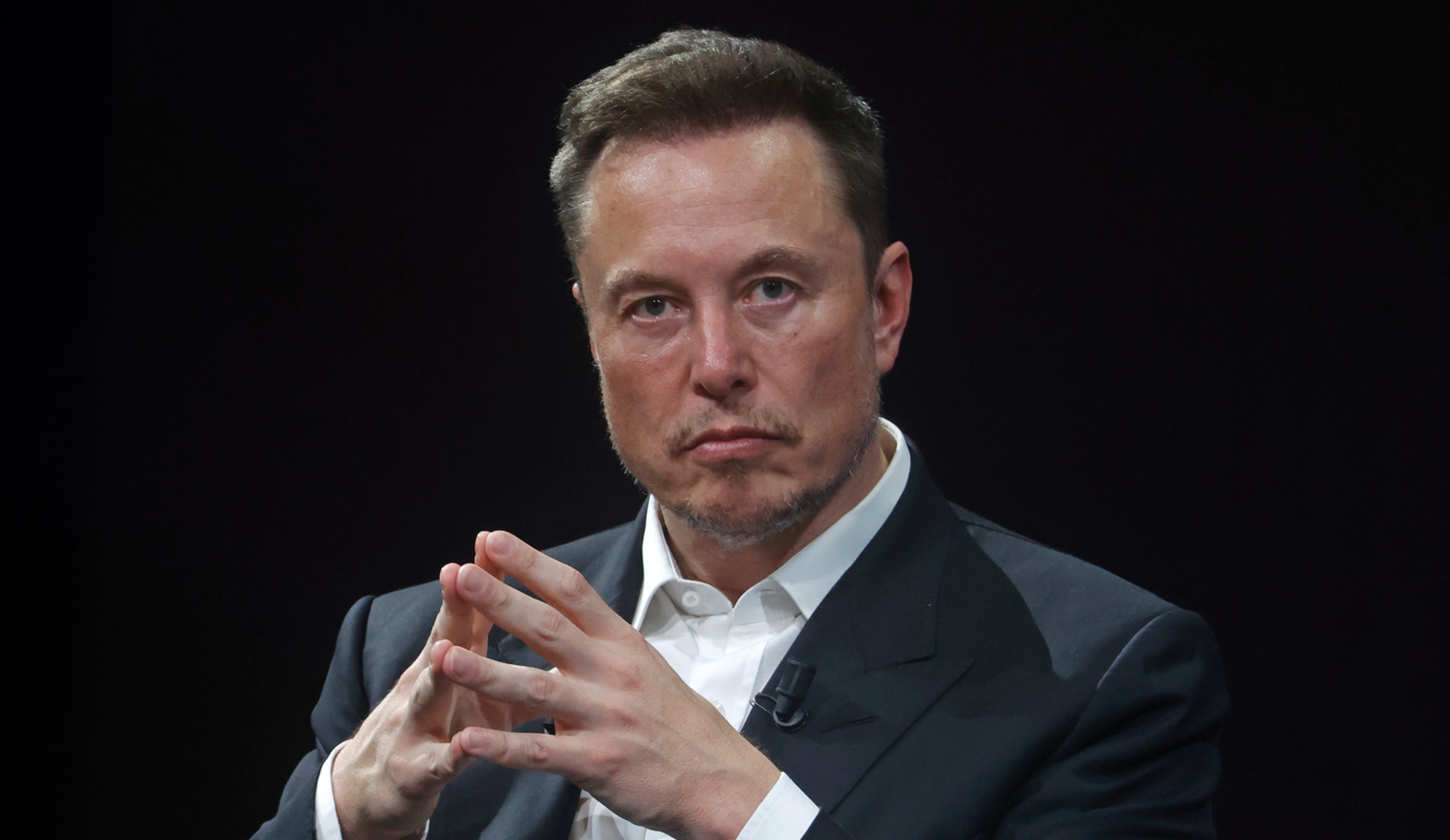 A man in a dark suit and white shirt sits with fingers interlaced, looking intently at the camera against a black background.