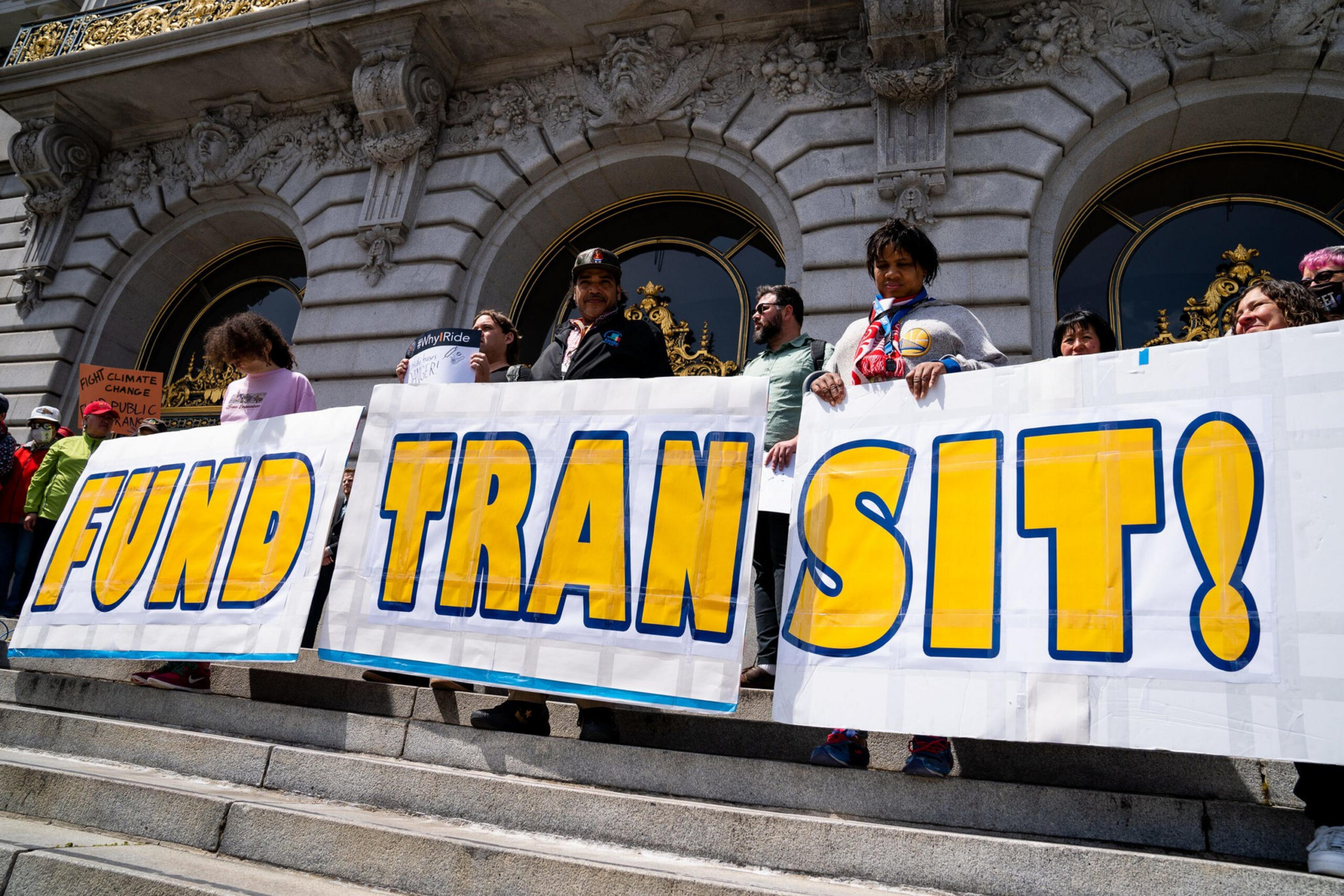 People stand on stone steps outside a building holding large signs that read "FUND TRANSIT!" in bold yellow and blue letters.