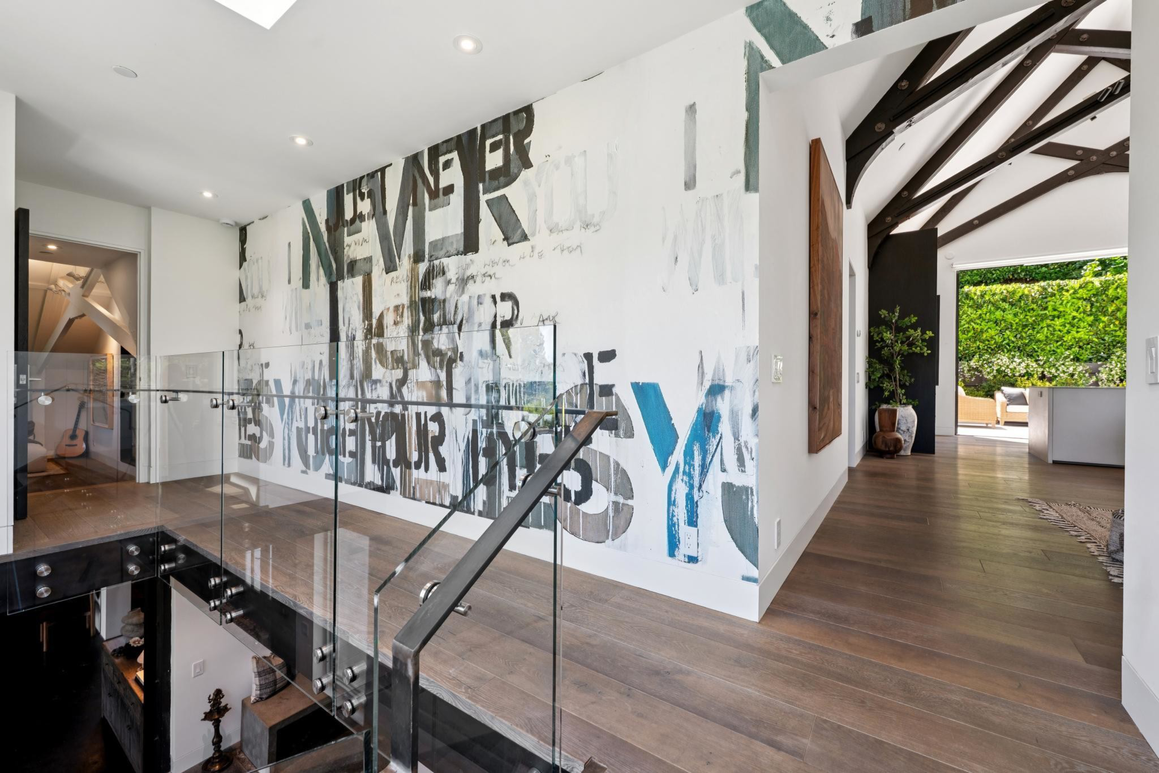 A modern hallway features wooden floors, a large abstract wall mural with bold, partially visible text, glass stair railings, and a view into a living area with greenery.