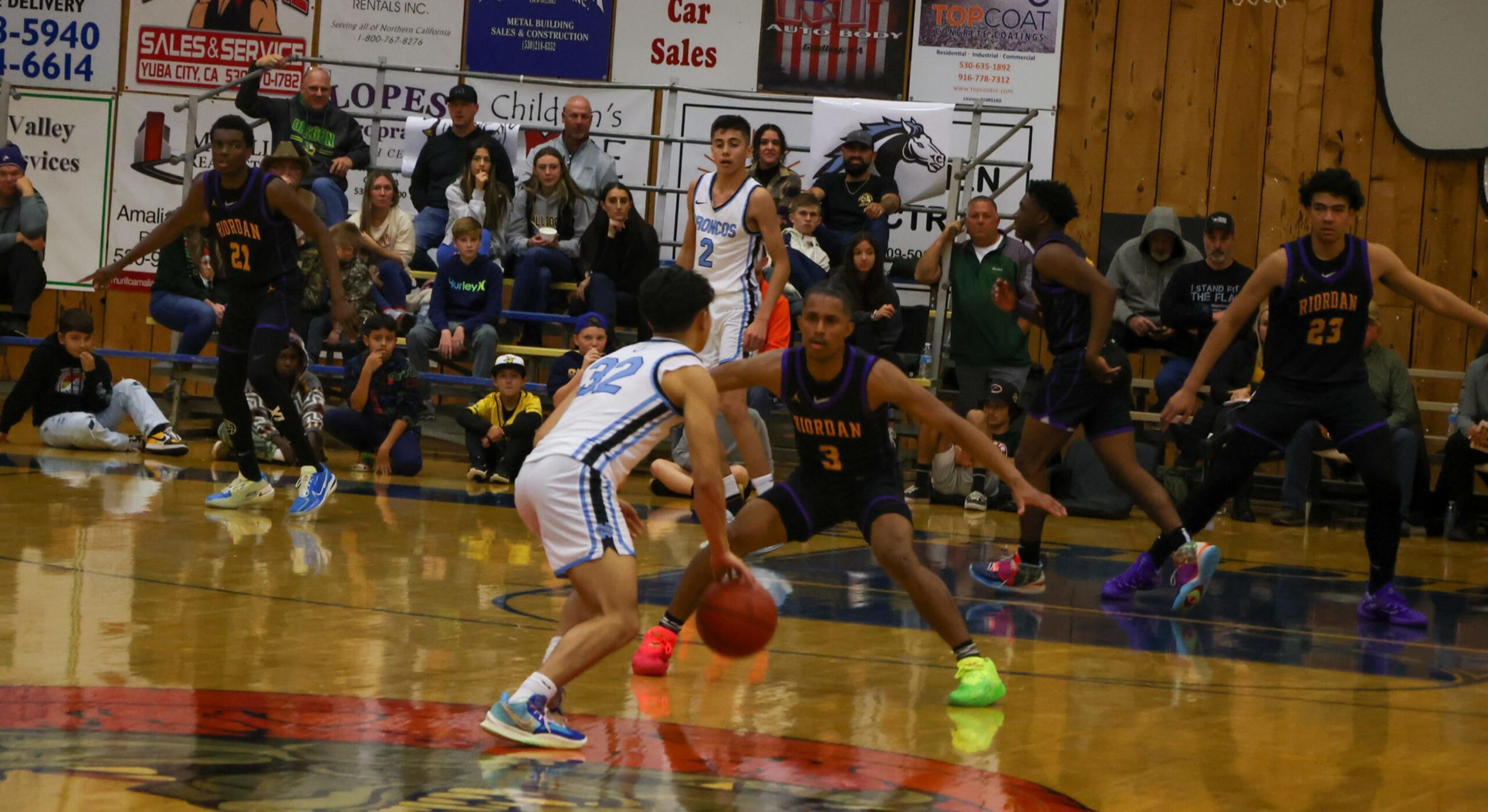 A basketball player in white dribbles the ball while three defenders in dark jerseys guard him, with spectators watching from the bleachers.