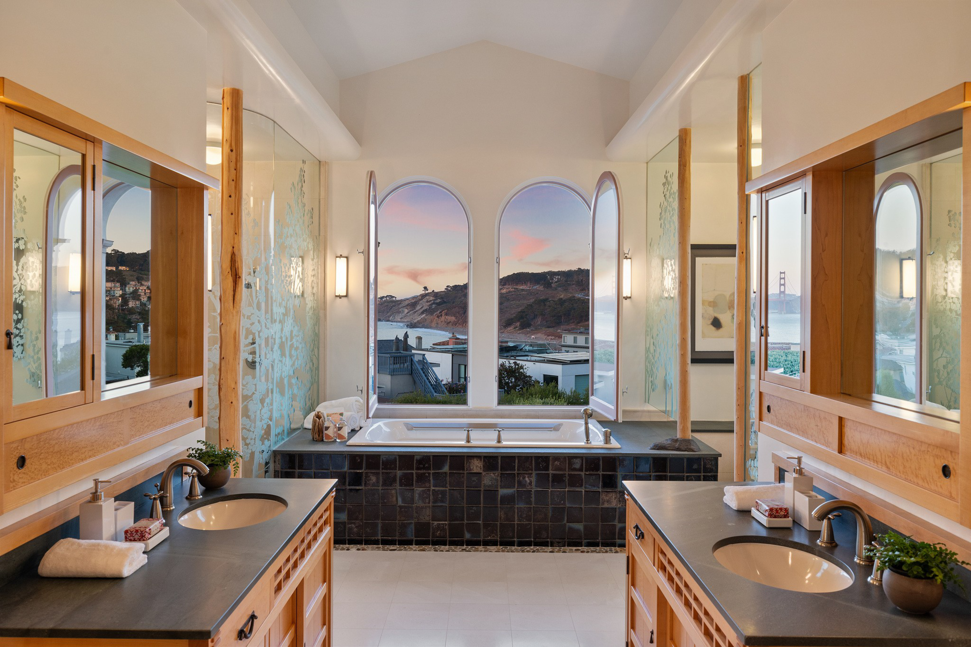 A spacious bathroom features a central bathtub with three arched windows overlooking hills and water, dual sinks with large mirrors, and natural wood accents.