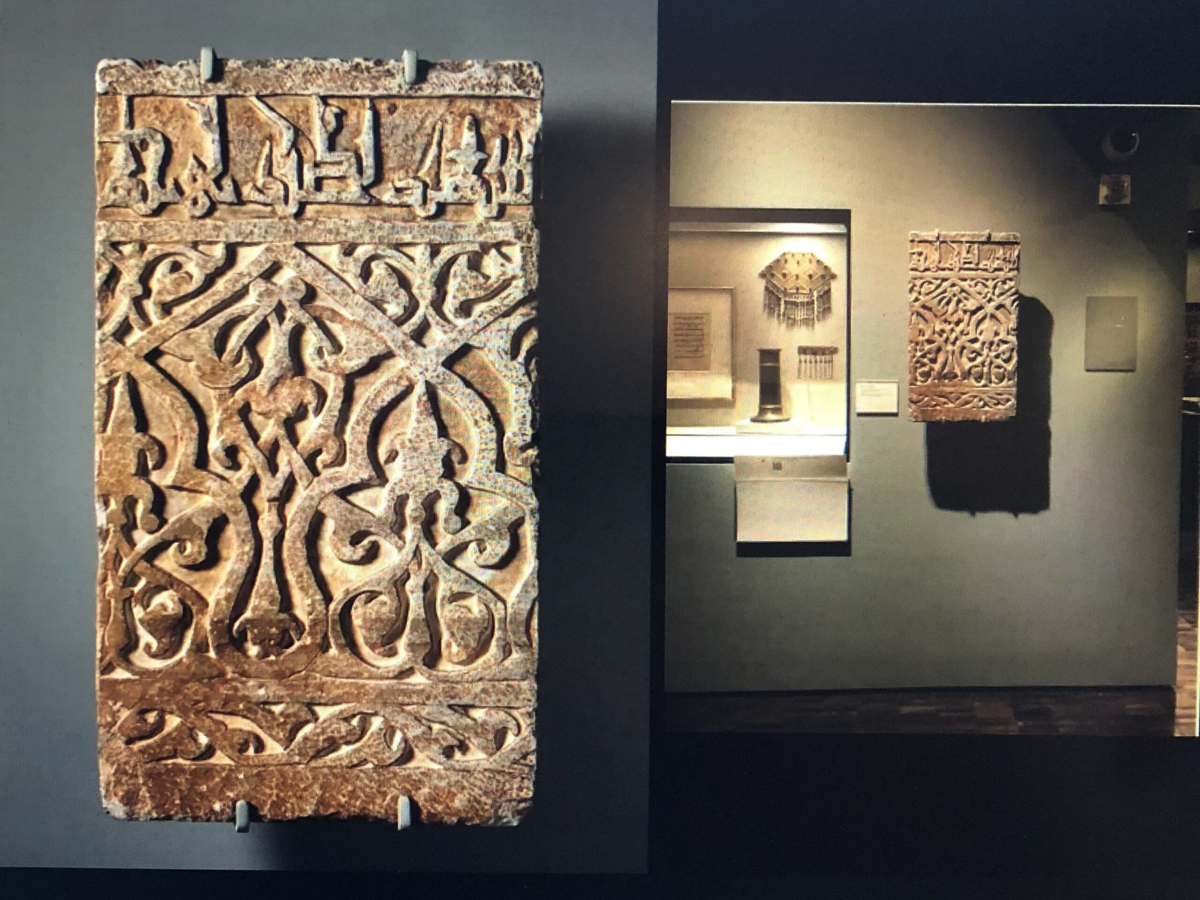 Ancient rectangular stone slab with intricate carved geometric and floral patterns, displayed on a dark wall in a museum setting.