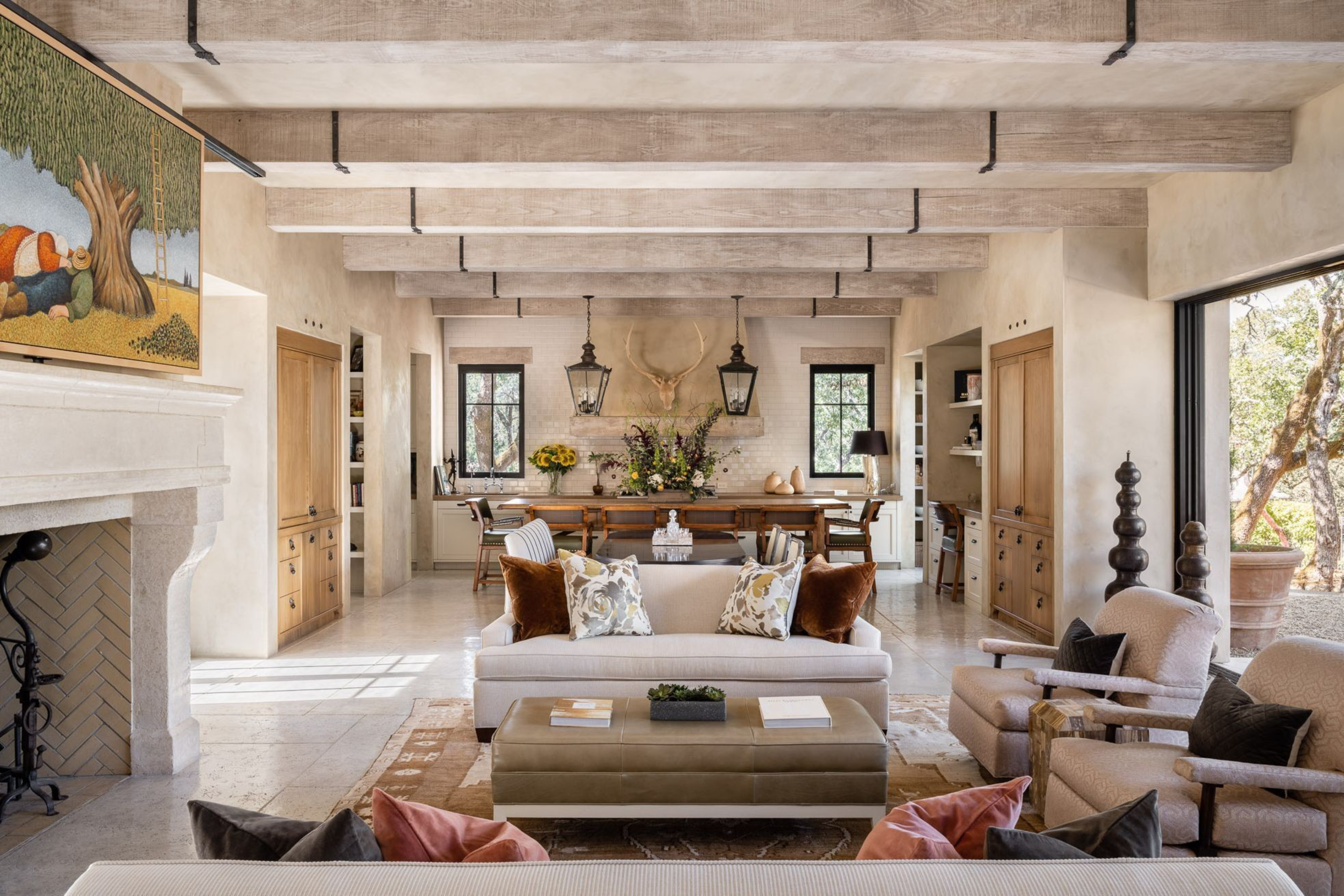 A cozy living room features a white sofa with patterned pillows, beige armchairs, a large fireplace with a colorful painting, wooden beams, and a dining area in the background.