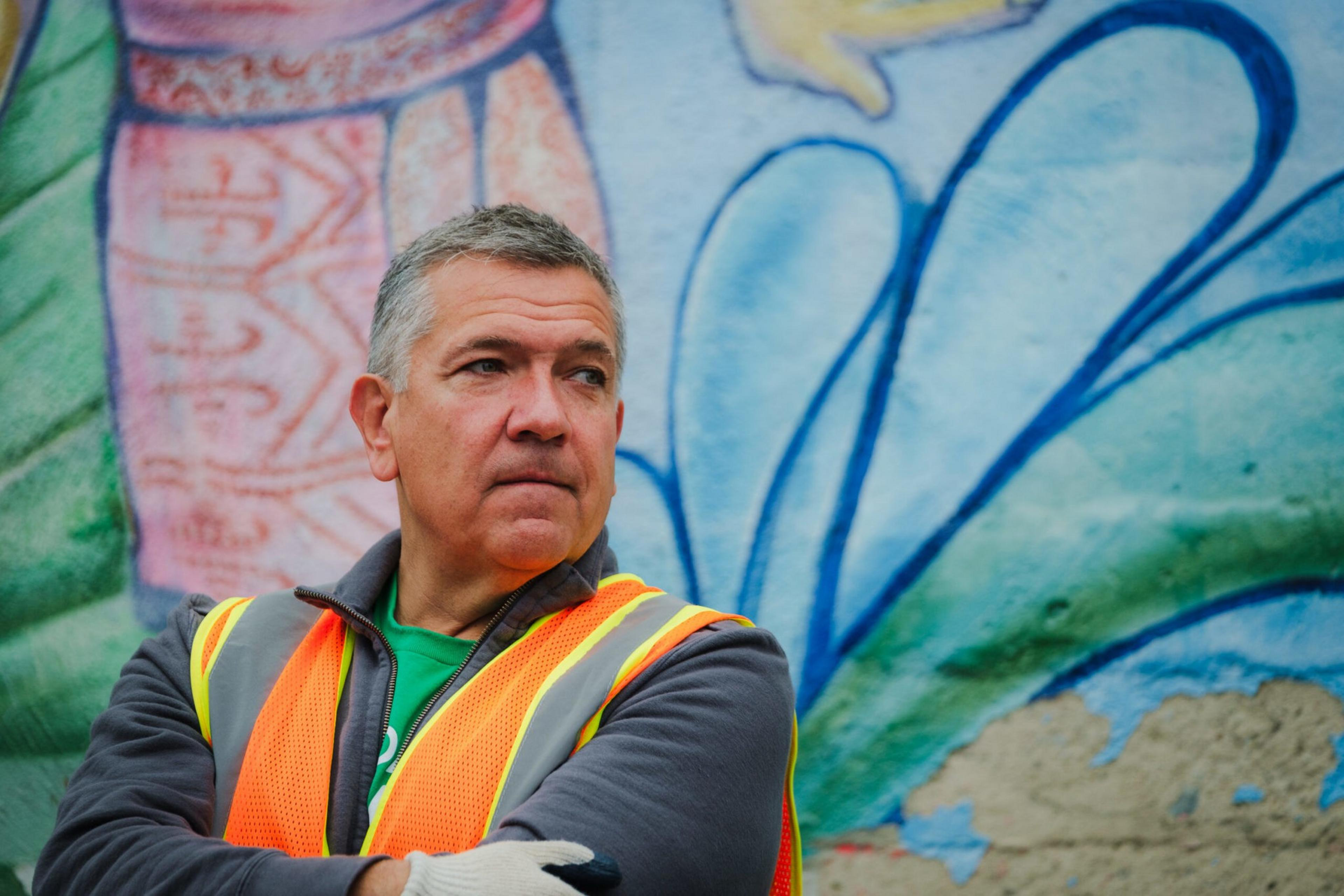 A middle-aged man in a neon orange safety vest and gray jacket stands with arms crossed in front of a colorful mural.
