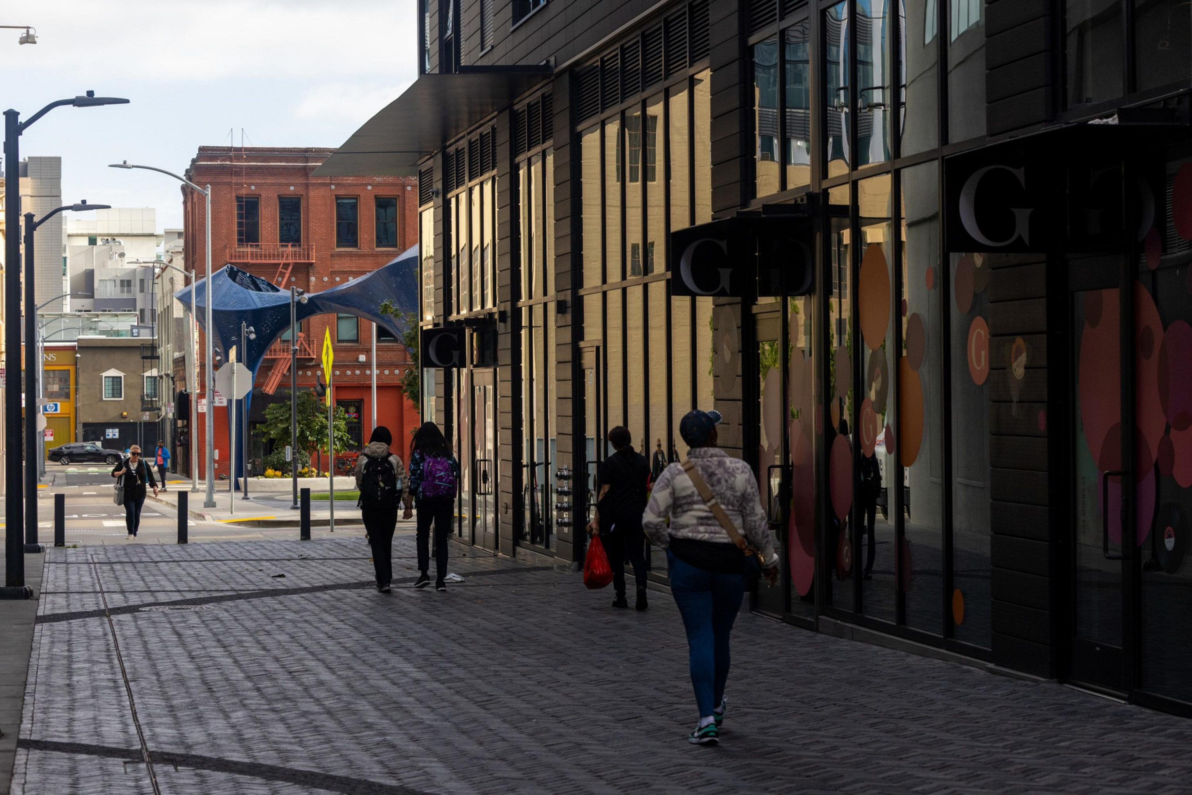People walk along a paved urban sidewalk next to a glass-fronted building with colorful circular designs, under a partly cloudy sky.
