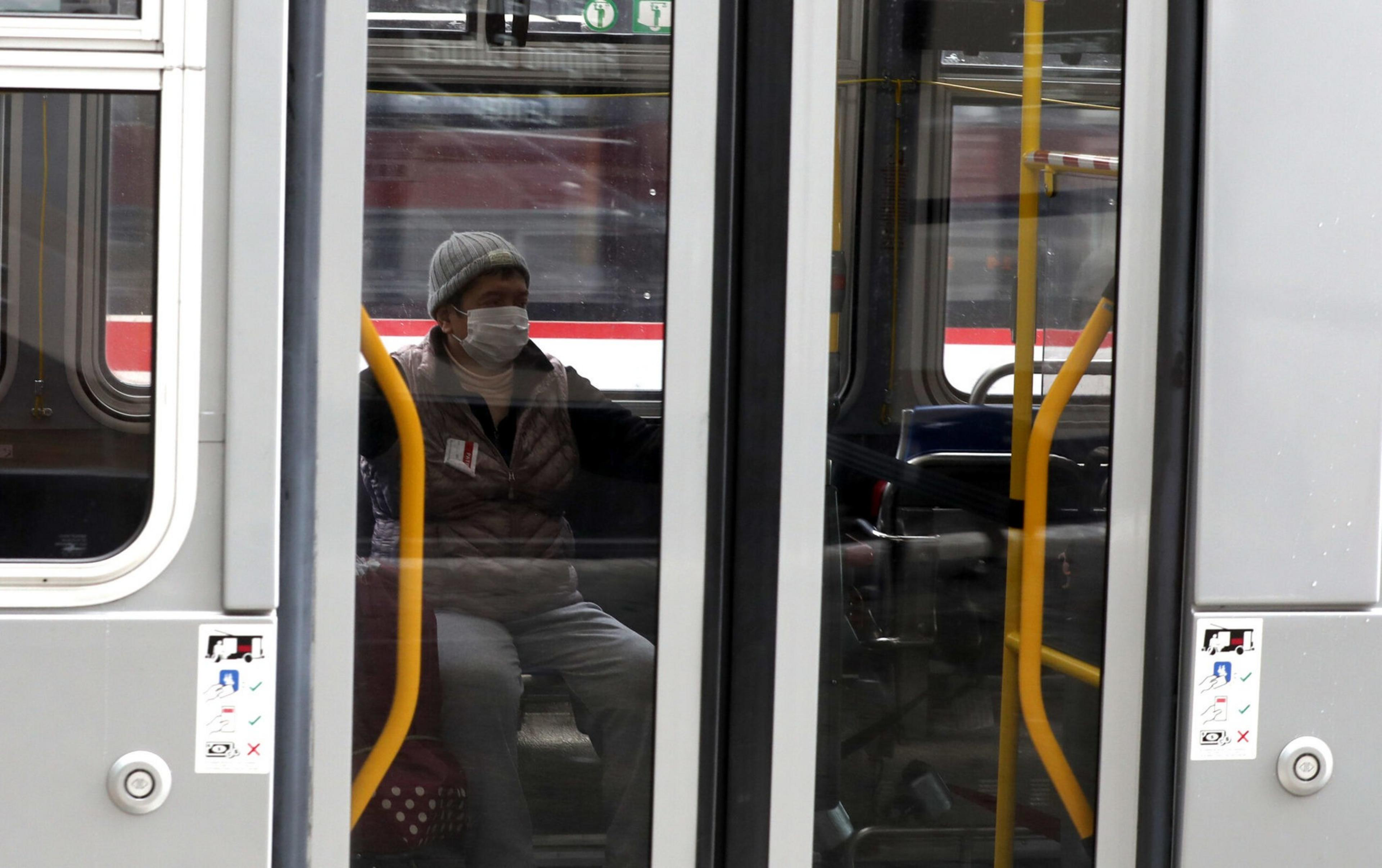 A person wearing a face mask and a beanie sits alone inside a public bus, visible through the closed doors with yellow handrails.