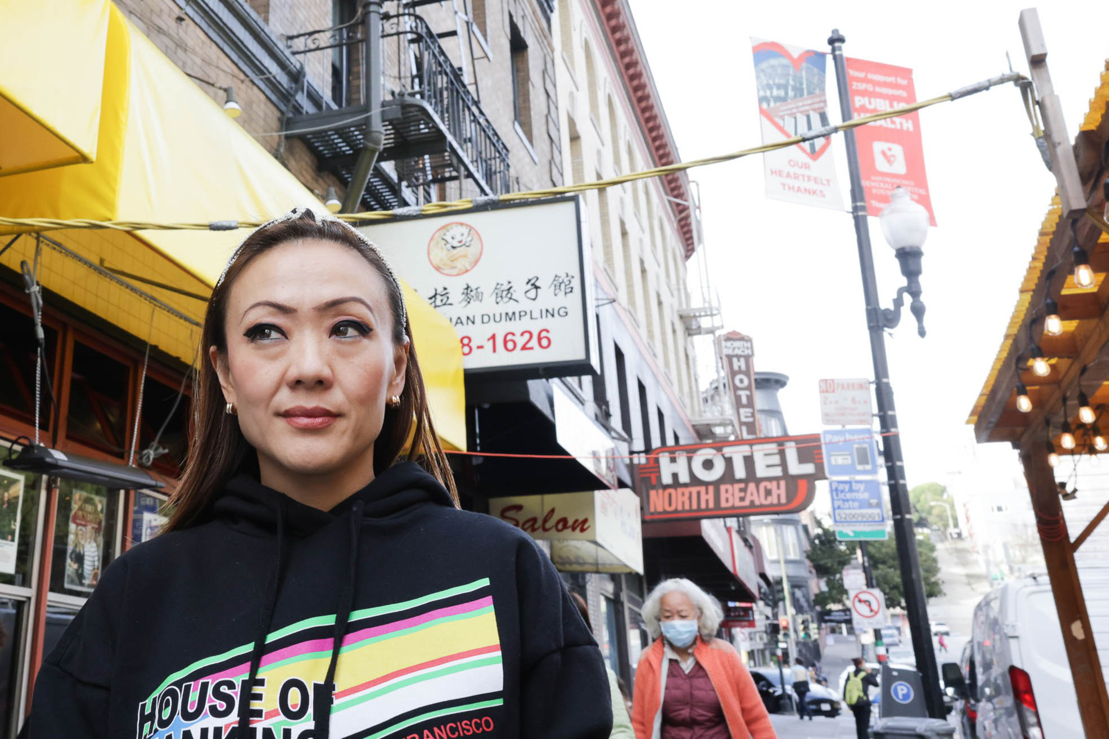 A woman in a hoodie stands on a city street with signs and lights in the background.