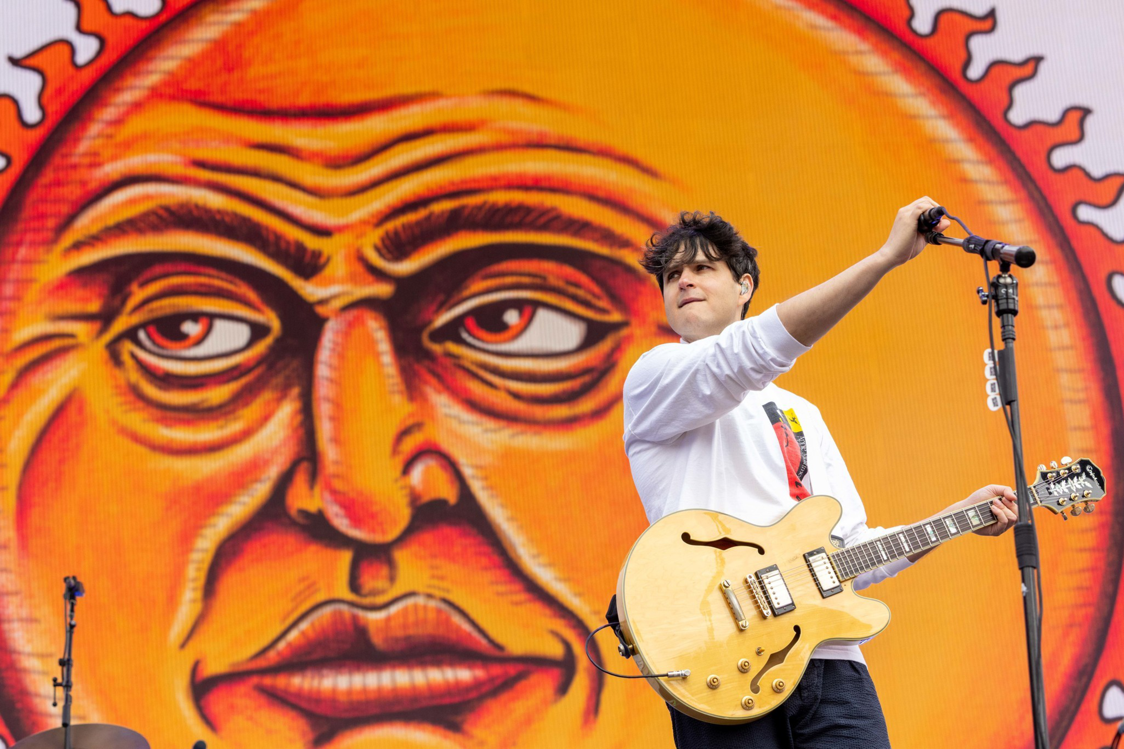 A musician with a yellow electric guitar adjusts a microphone, standing in front of a large, detailed orange sun face backdrop.