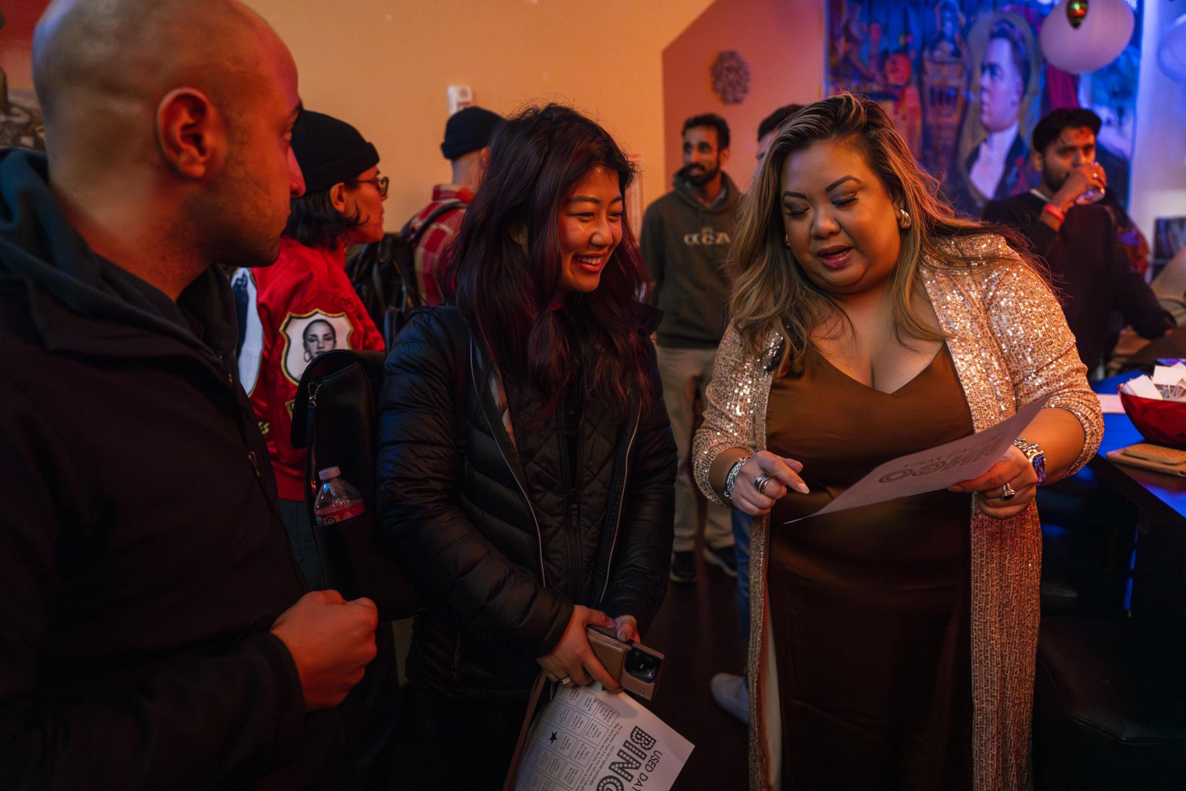 A woman shows a party attendee how to play &quot;Used Date Party&quot; bingo at a single mixers event in a low lit bar in San Francisco.