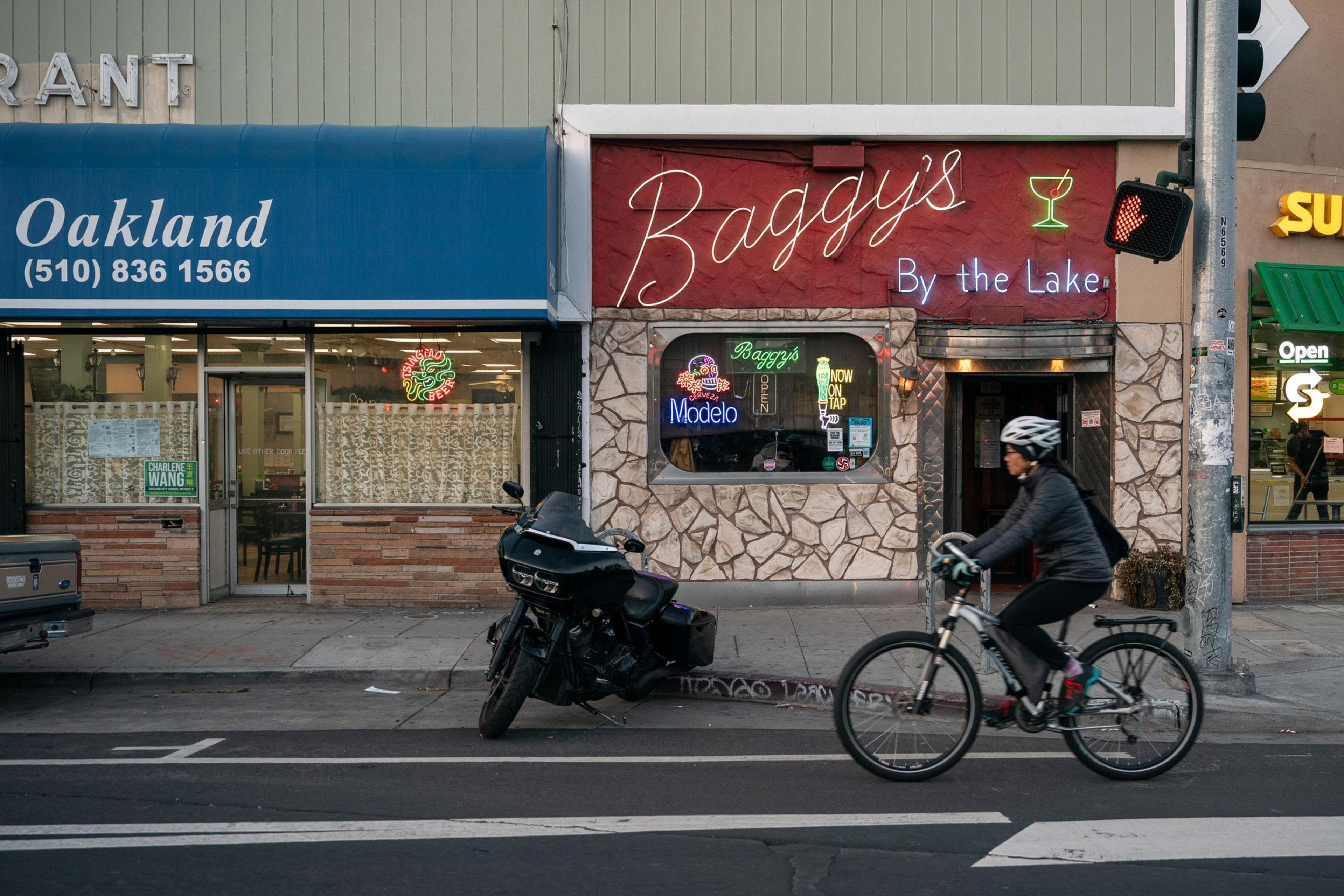 A cyclist rides past a row of storefronts including Baggy's Bar and an Oakland restaurant. A motorcycle is parked on the sidewalk beside the bar.
