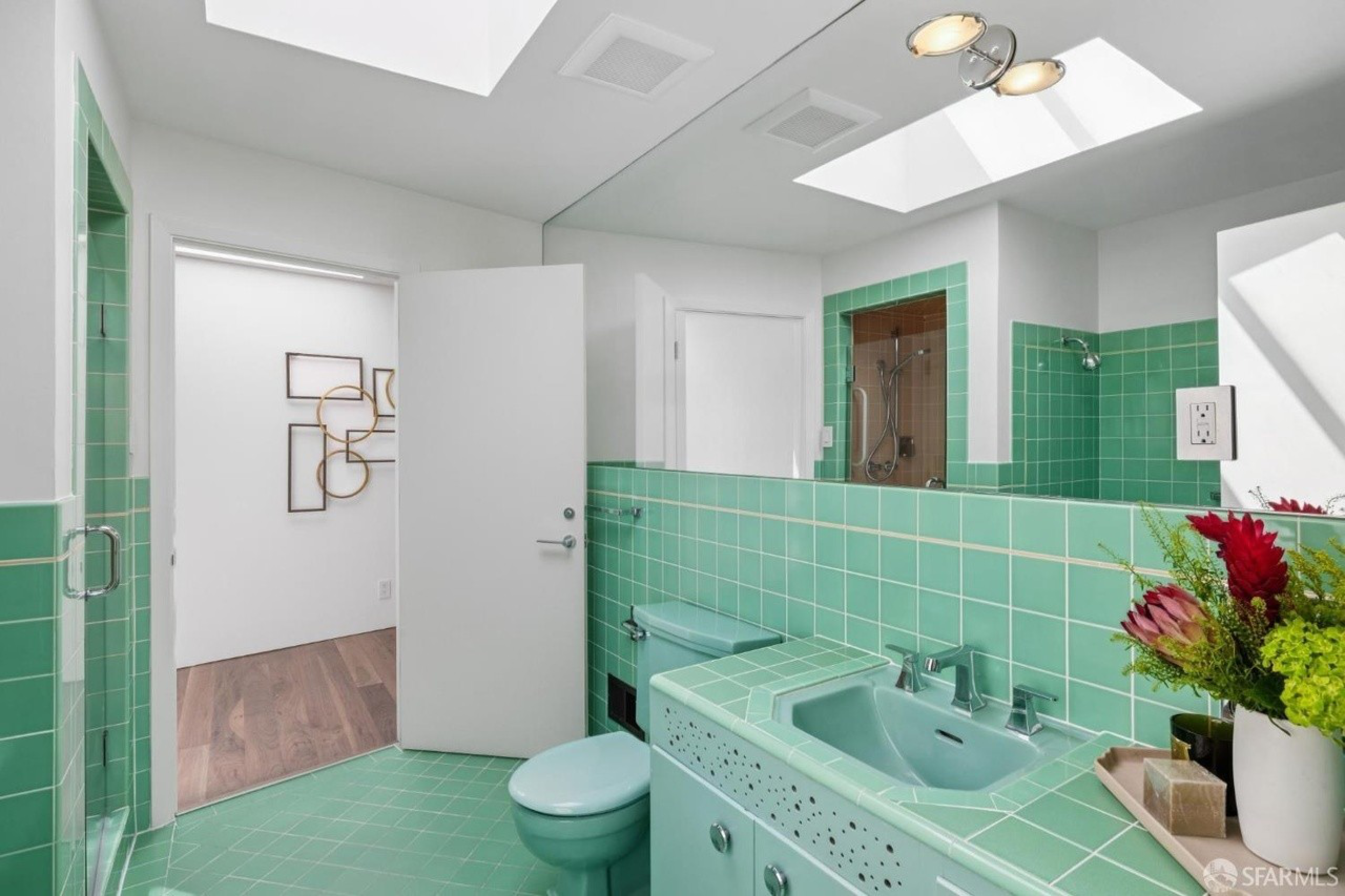 A bathroom with green tiled walls and floor, a green toilet, a green sink, a large mirror, skylights, and a vase with colorful flowers on the counter.