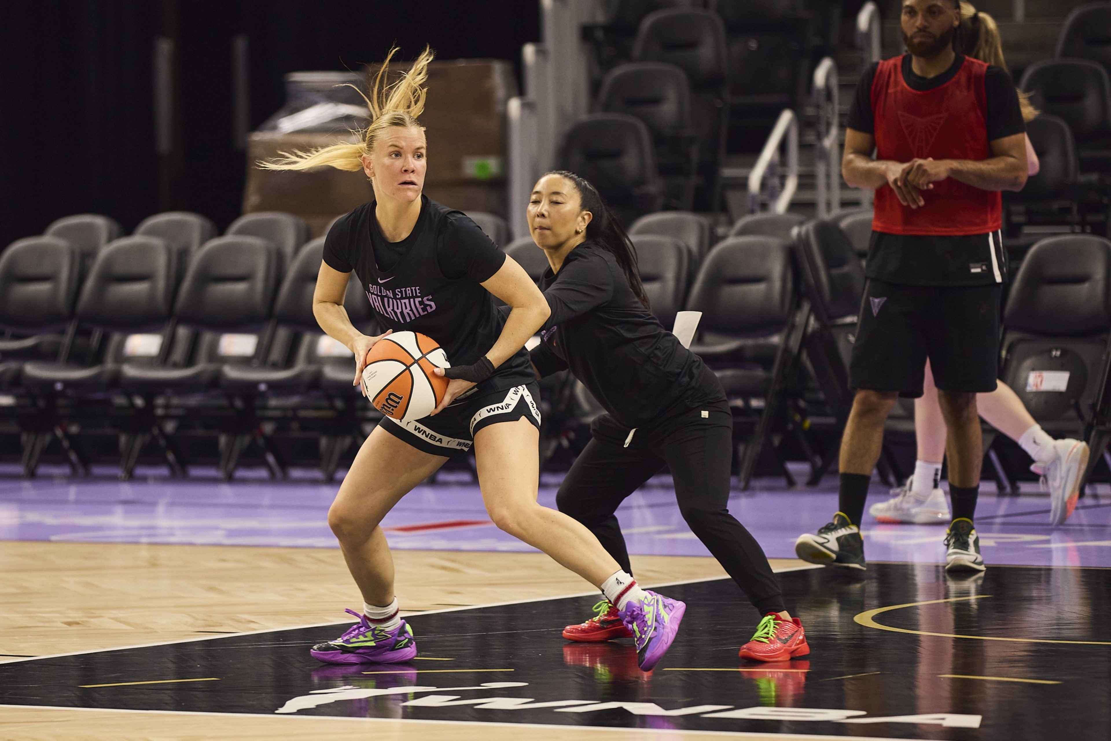 A basketball player in a &quot;Golden State Valkyries&quot; shirt is intensely dribbling with a defender close by. Others watch from the background on an indoor court.
