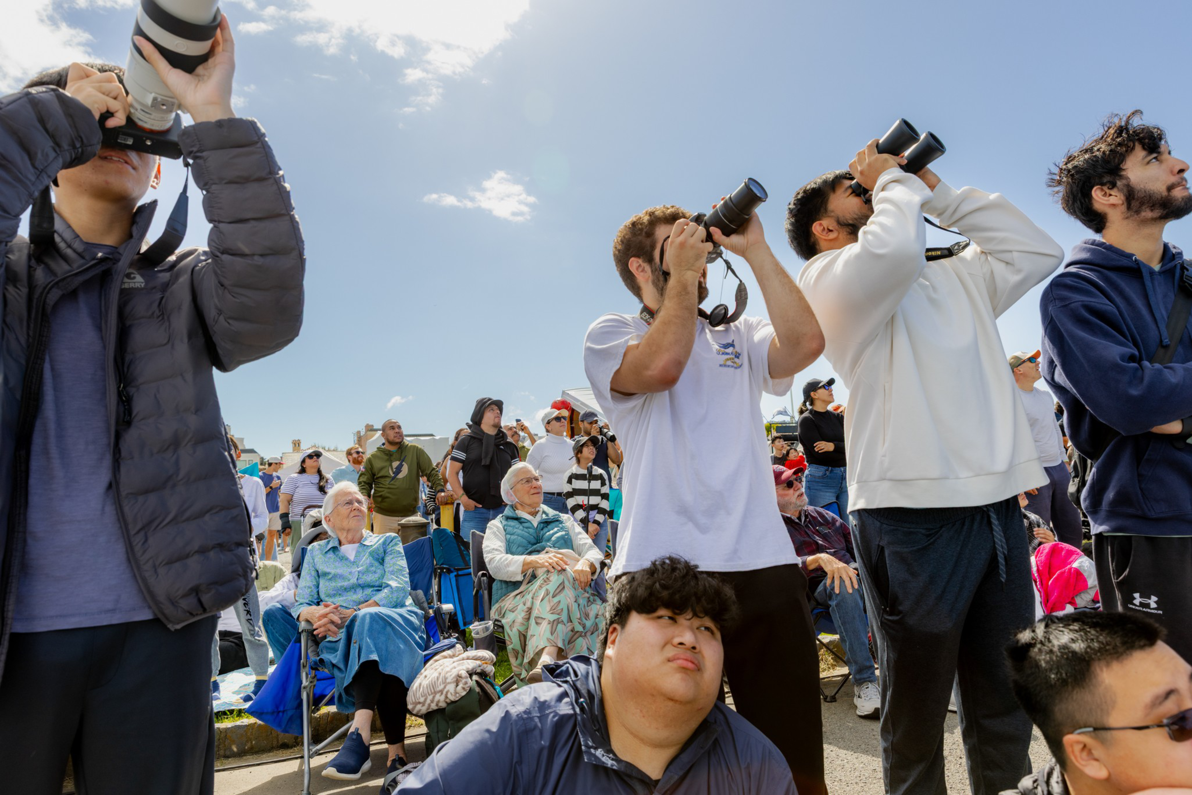 A diverse crowd of people including photographers with cameras and binoculars looks up at the sky on a sunny day.