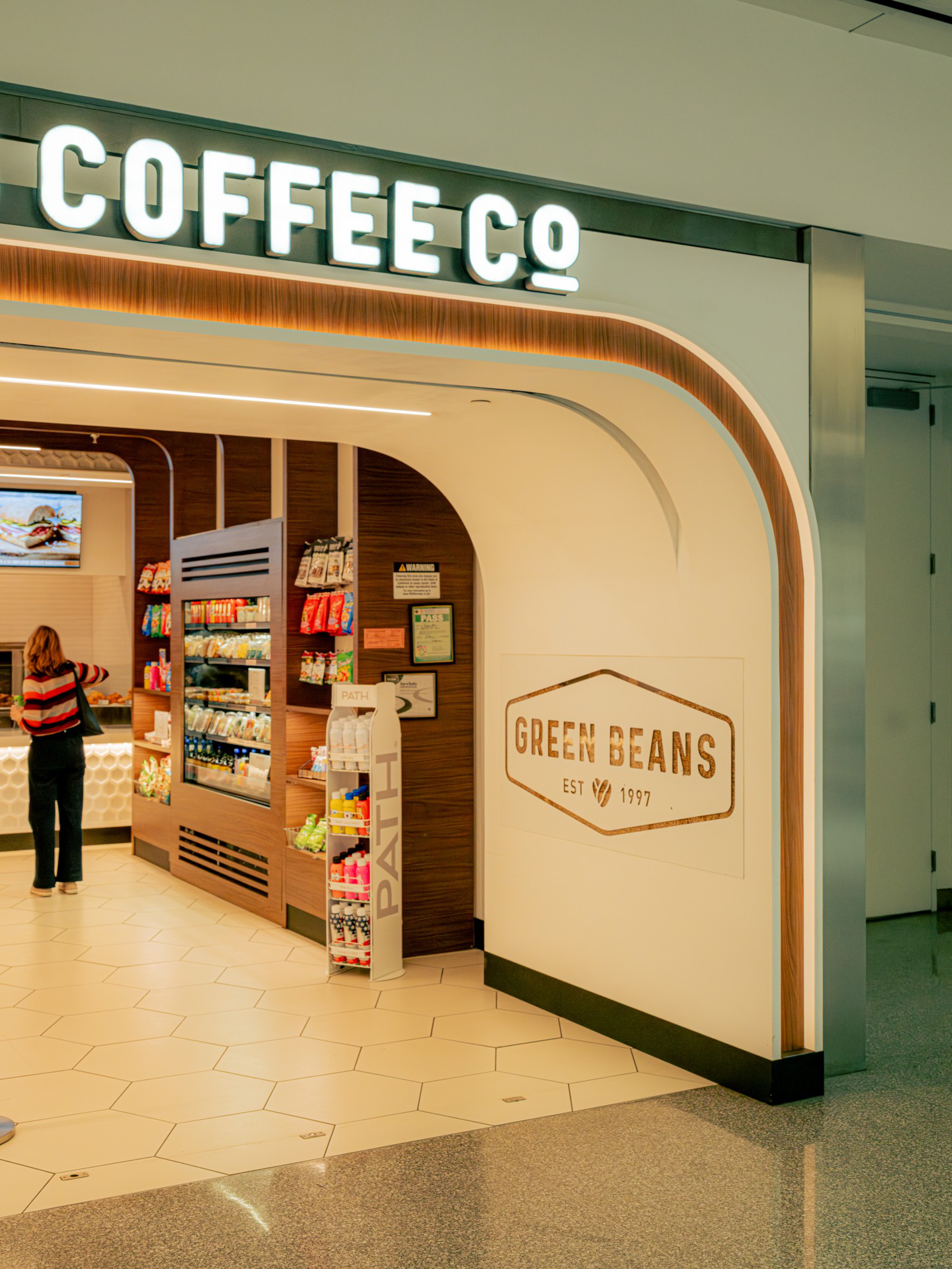 A modern coffee shop named Green Beans with a bright “COFFEE CO” sign, wooden shelves stocked with snacks, and a customer inside.