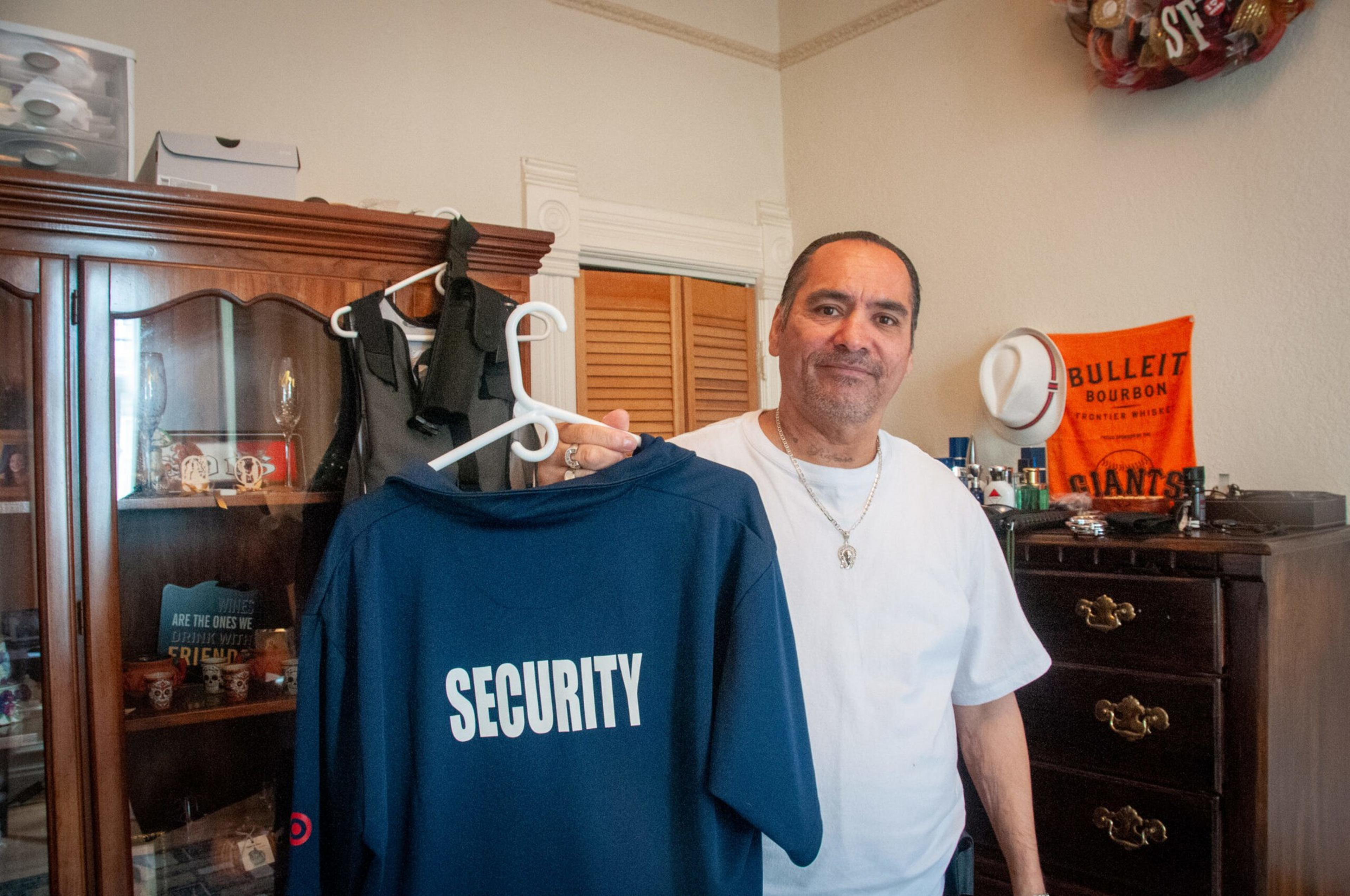 A man in a white shirt holds up a navy blue shirt with "SECURITY" printed on the back inside a room with wooden furniture and personal items.