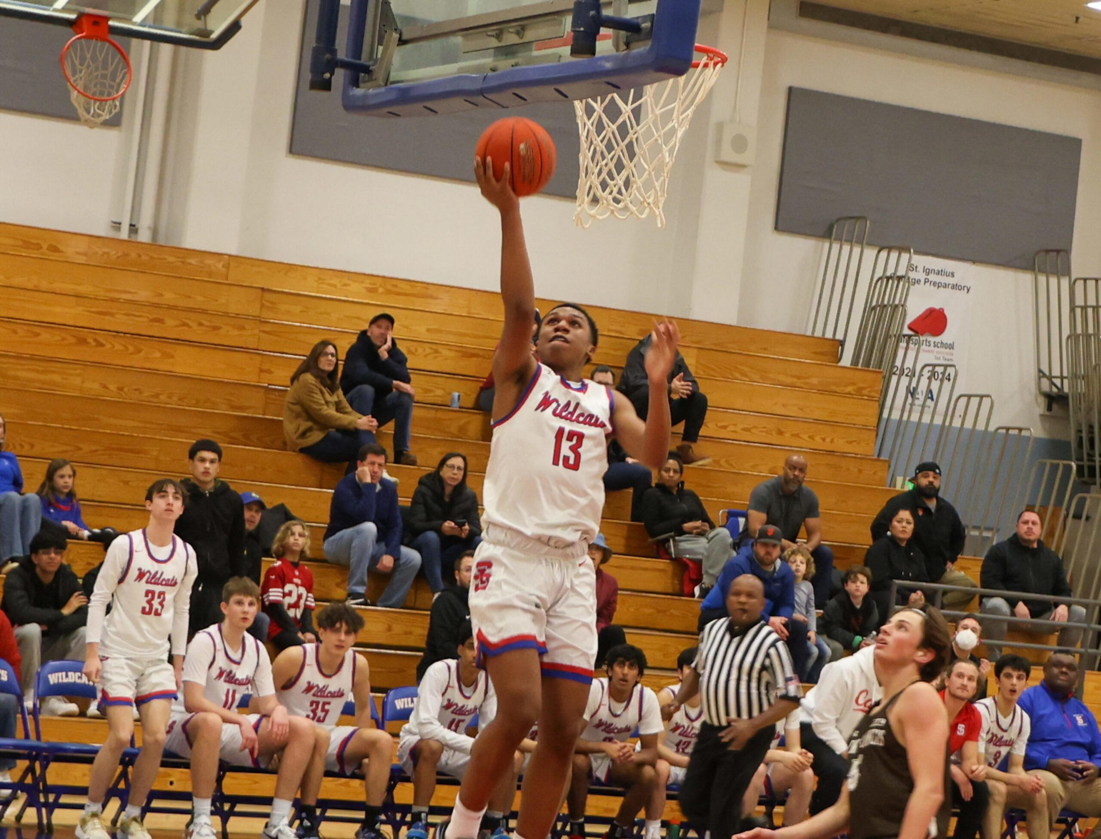 A basketball player wearing a white Wildcats jersey with number 13 is jumping to make a layup while spectators and teammates watch.
