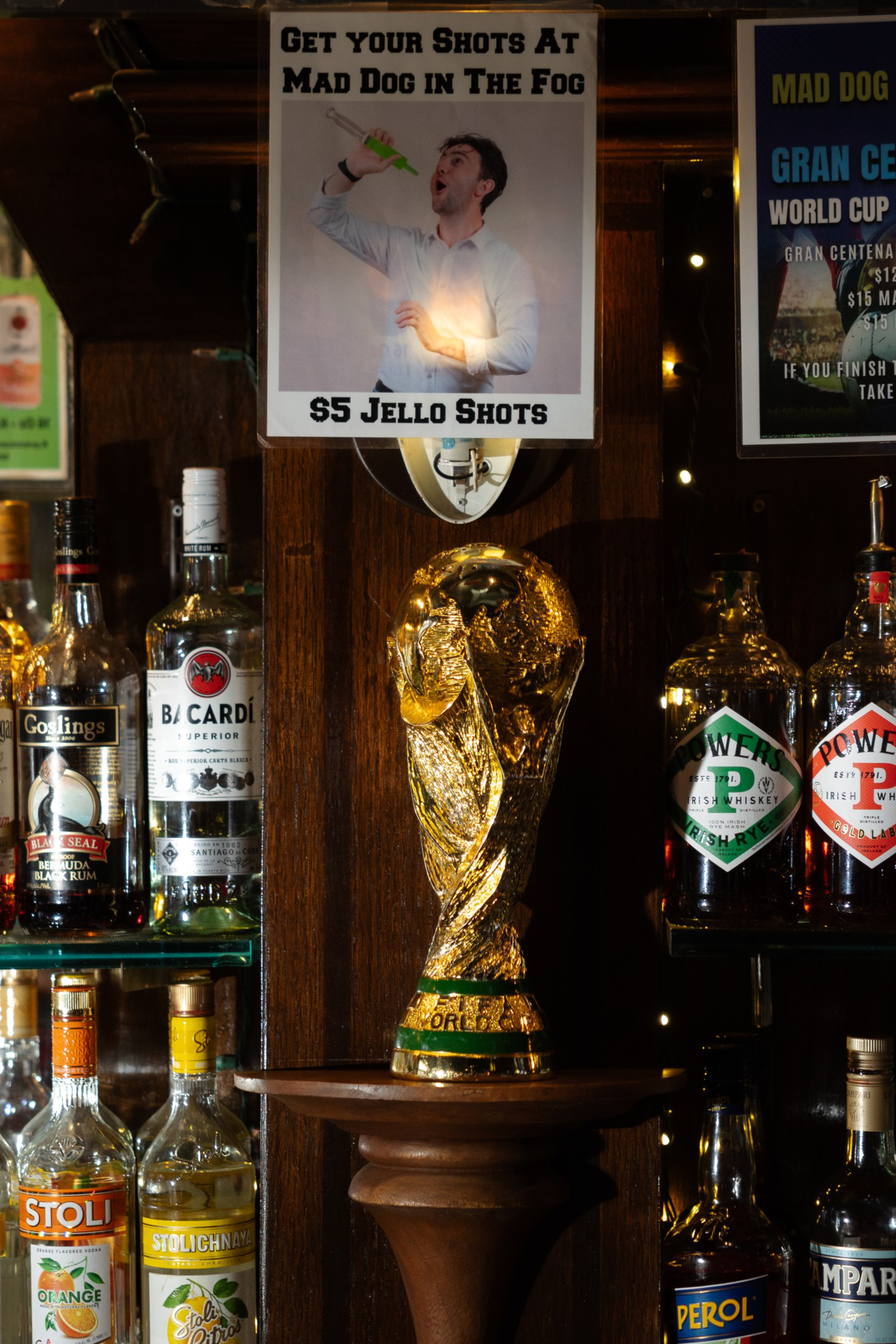 A golden trophy resembling the FIFA World Cup sits on a wooden pedestal, surrounded by various liquor bottles and a sign advertising $5 Jello shots.