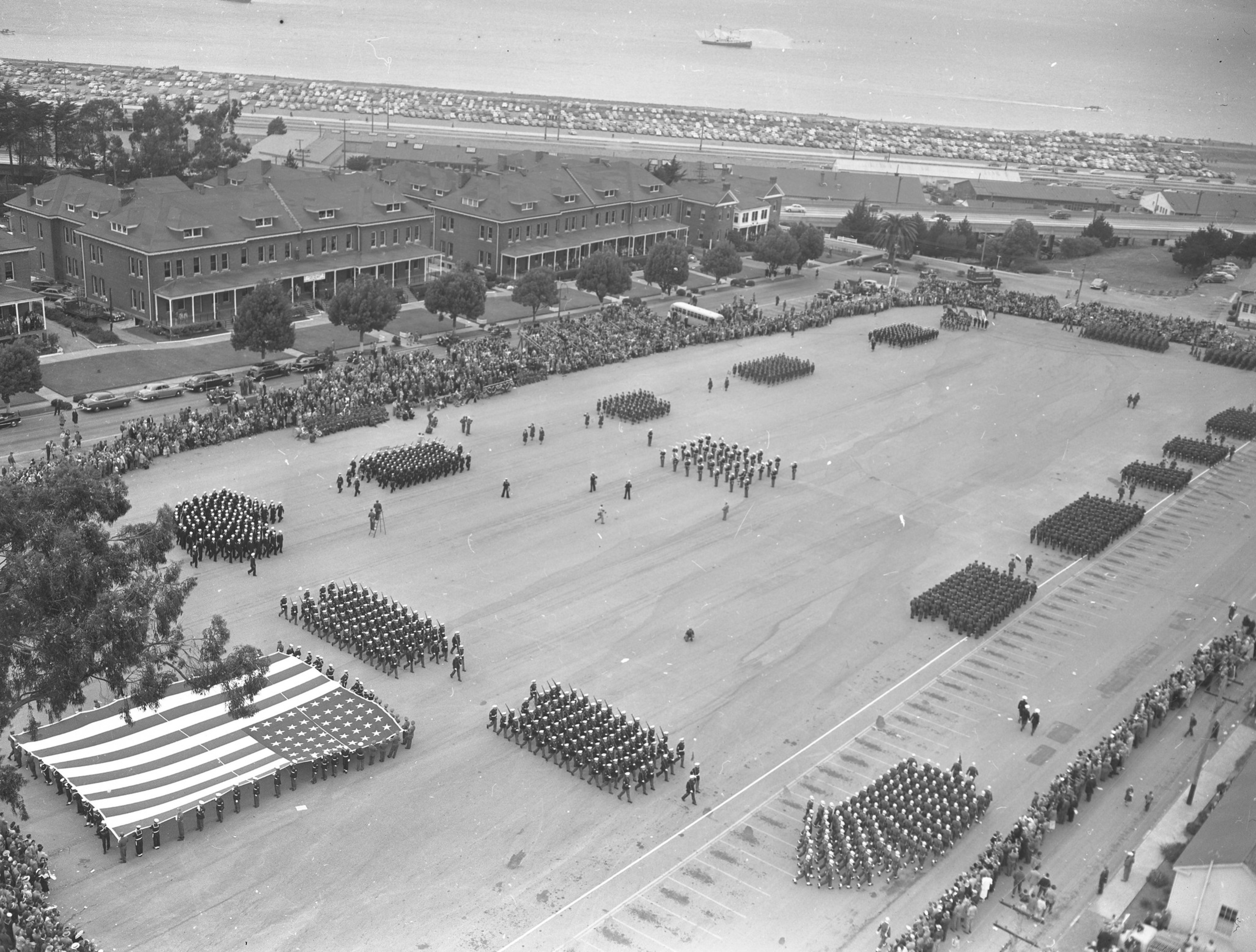 Military parade in a wide field near water, with formations of troops and spectators.