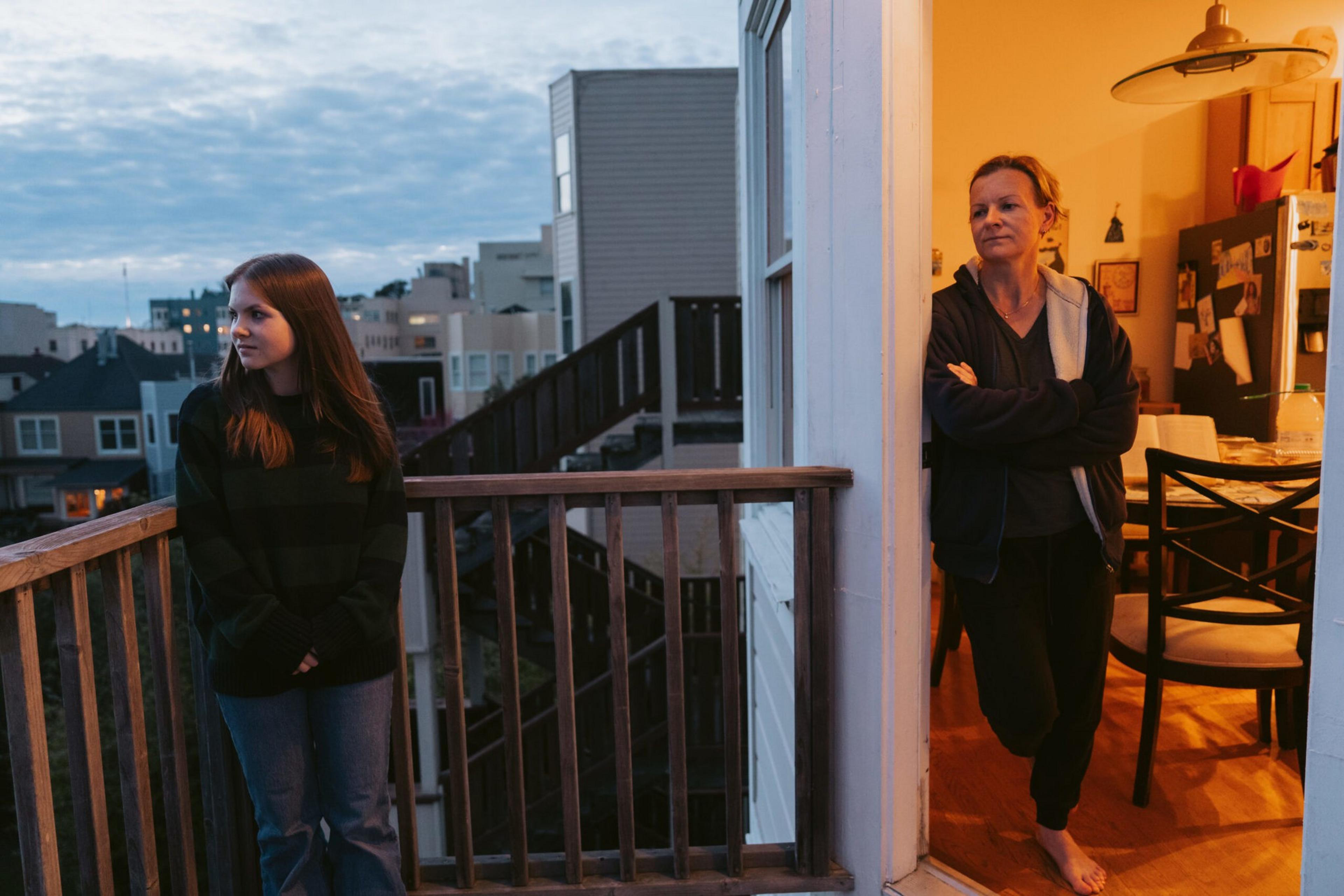 A teenage girl stands outside on a balcony at dusk, looking away while a woman stands inside near the doorway with arms crossed, both appearing thoughtful.