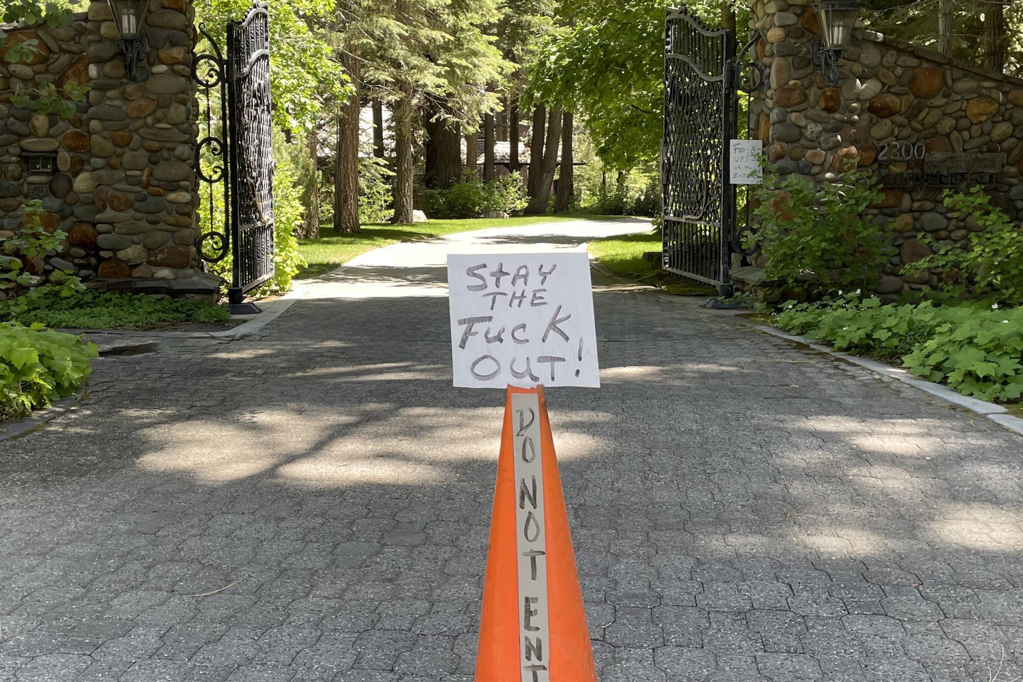 The image shows a gated driveway with a stone entrance, and a traffic cone bearing a sign that reads, "STAY THE FUCK OUT!" with "DO NOT ENTER" written vertically on the cone.