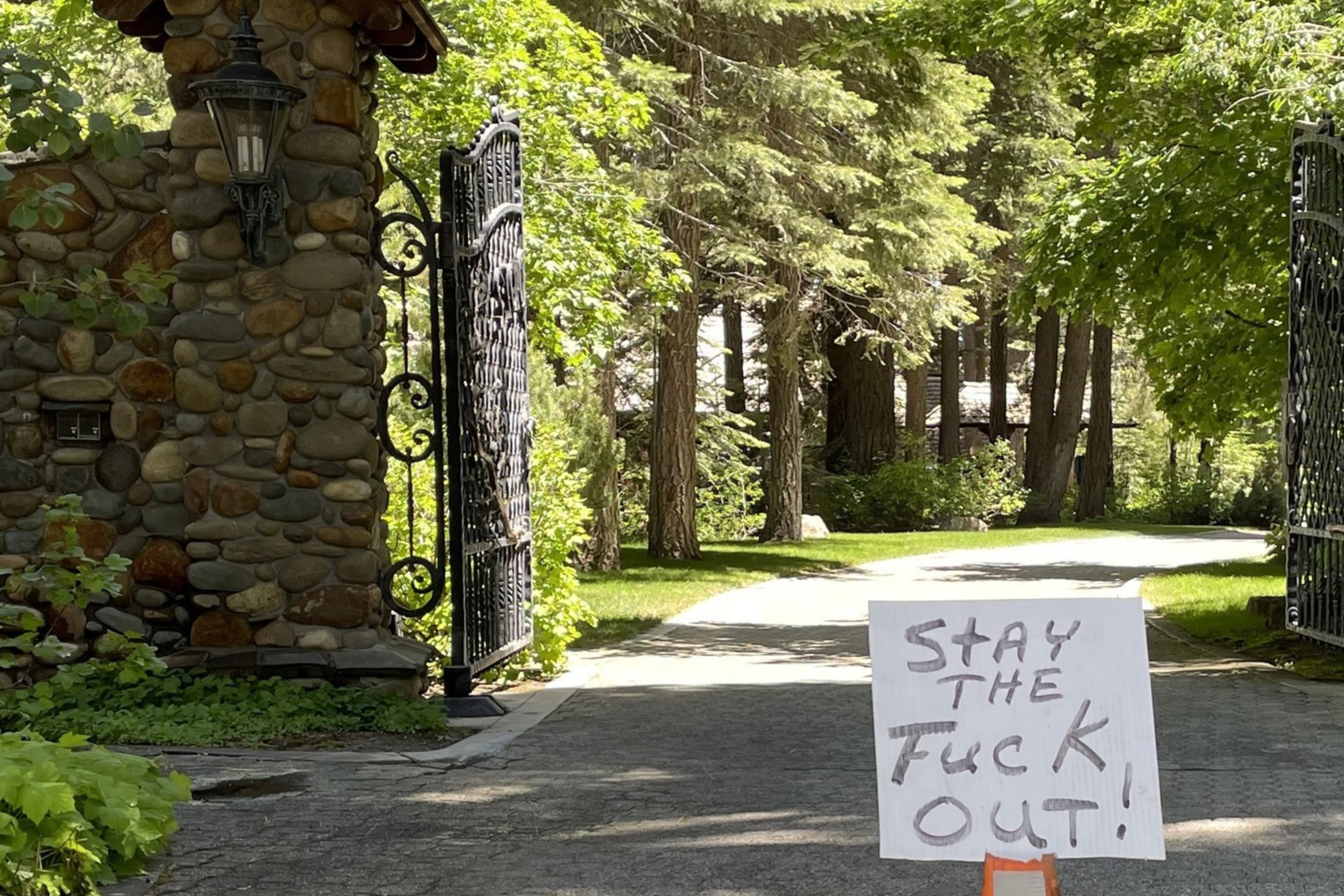 The image shows a gated driveway with a stone entrance, and a traffic cone bearing a sign that reads, "STAY THE FUCK OUT!" with "DO NOT ENTER" written vertically on the cone.