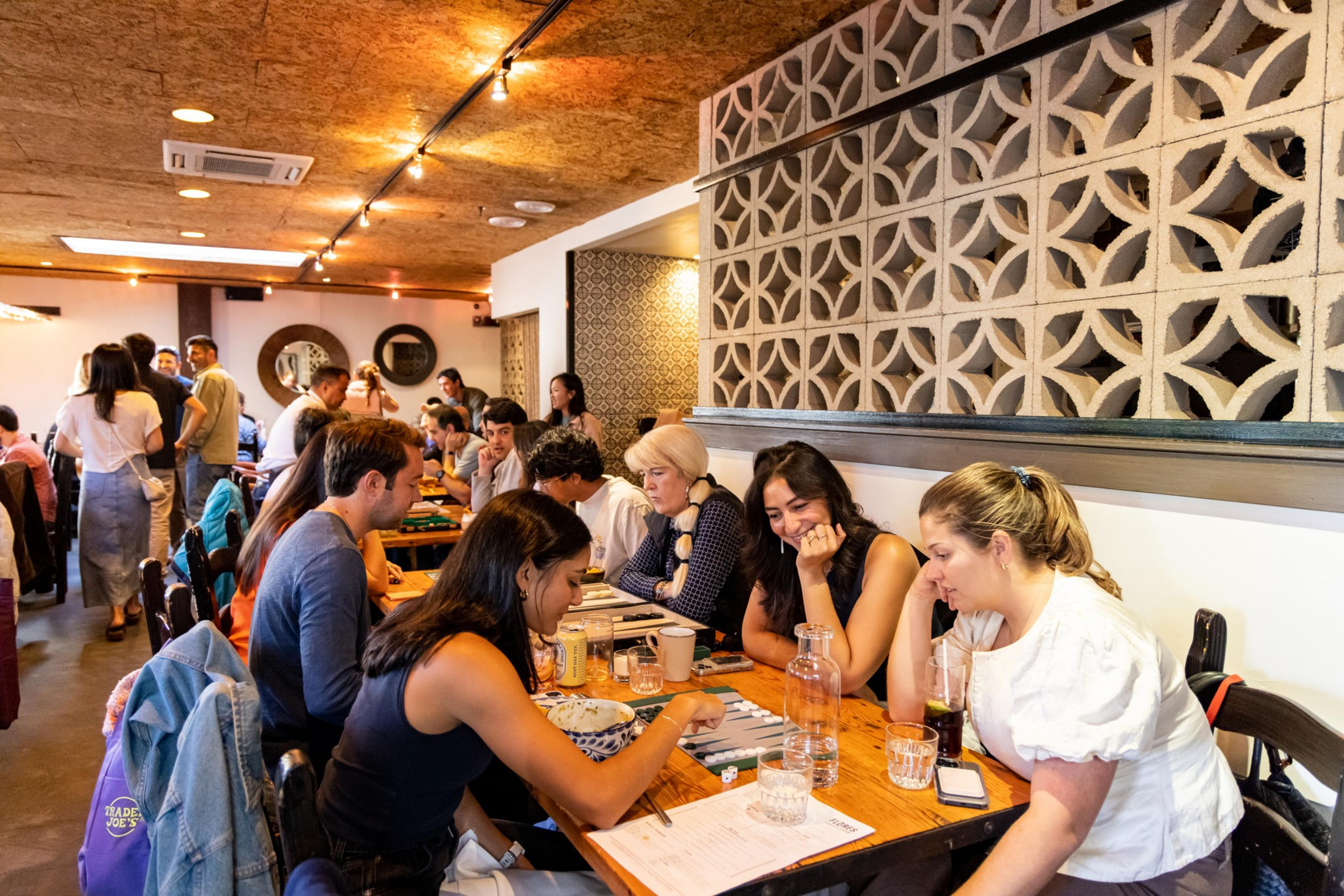 People are seated closely at a long wooden table in a lively café, engaged in conversations and playing board games.