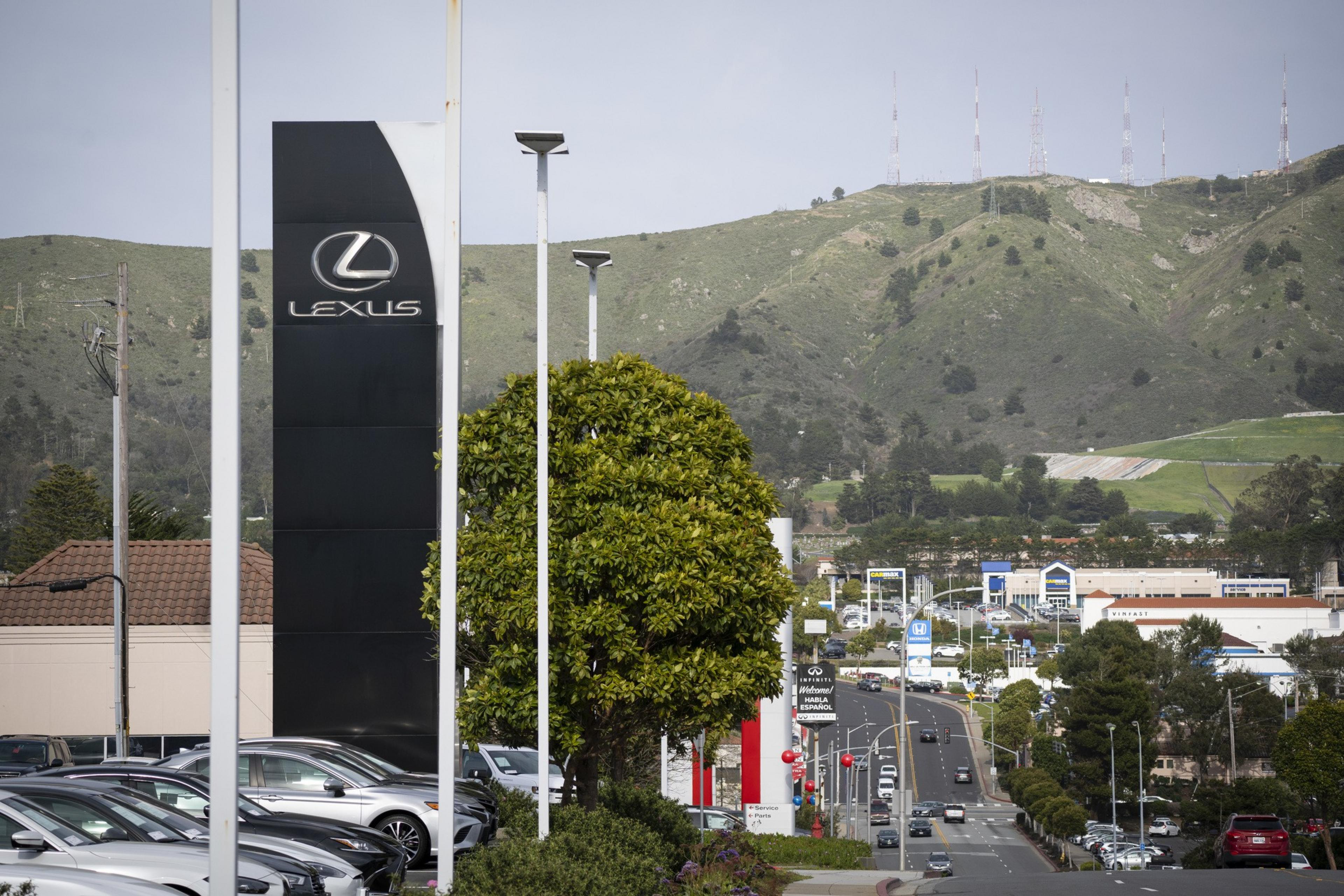 A Lexus dealership sign stands beside parked cars, with a tree and streetlights nearby. In the background, a road leads toward hills and transmission towers.