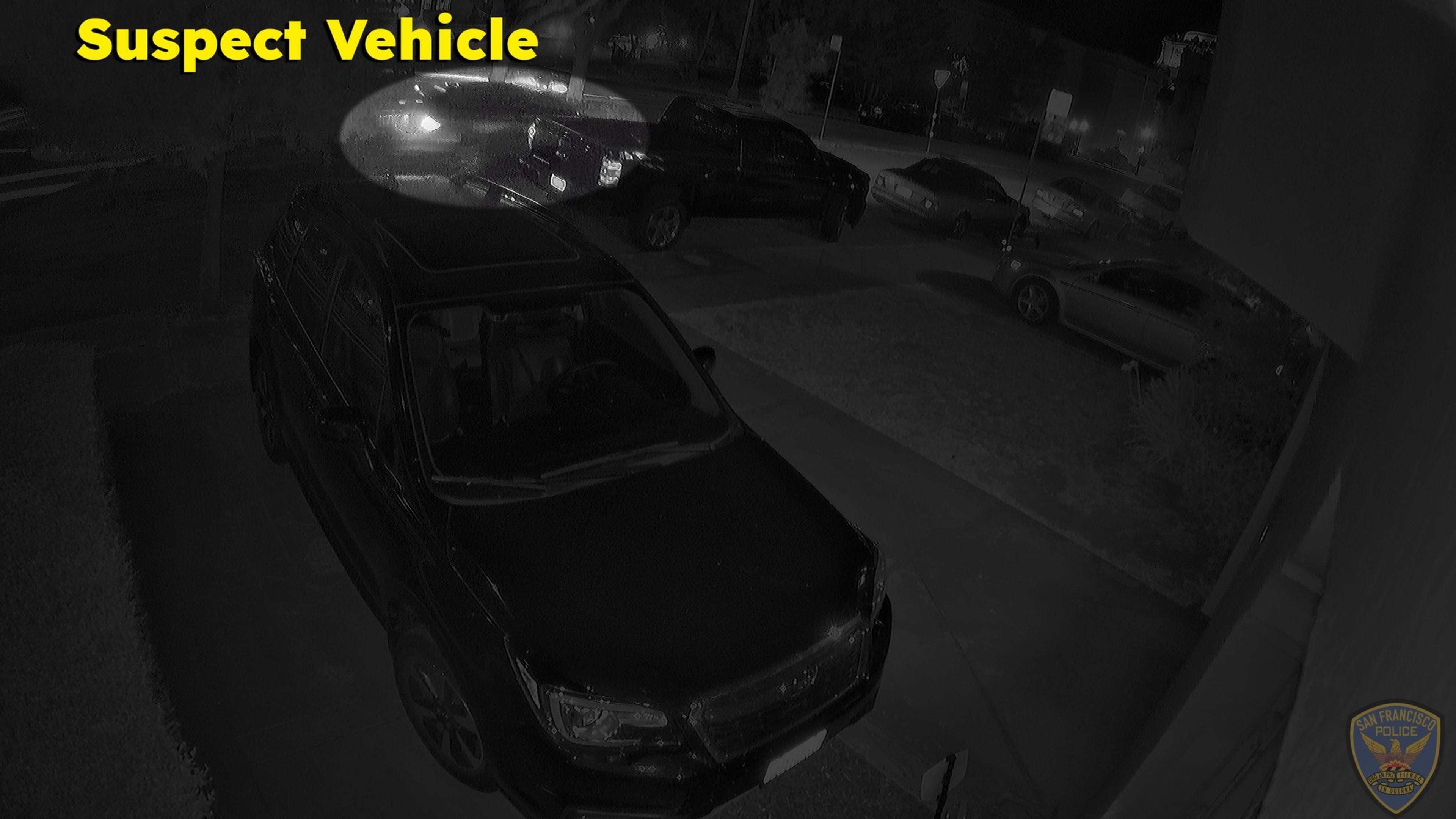 A dark SUV is parked in a driveway at night, with several cars parked along the street nearby under dim lighting. The words "Suspect Vehicle" appear in yellow.