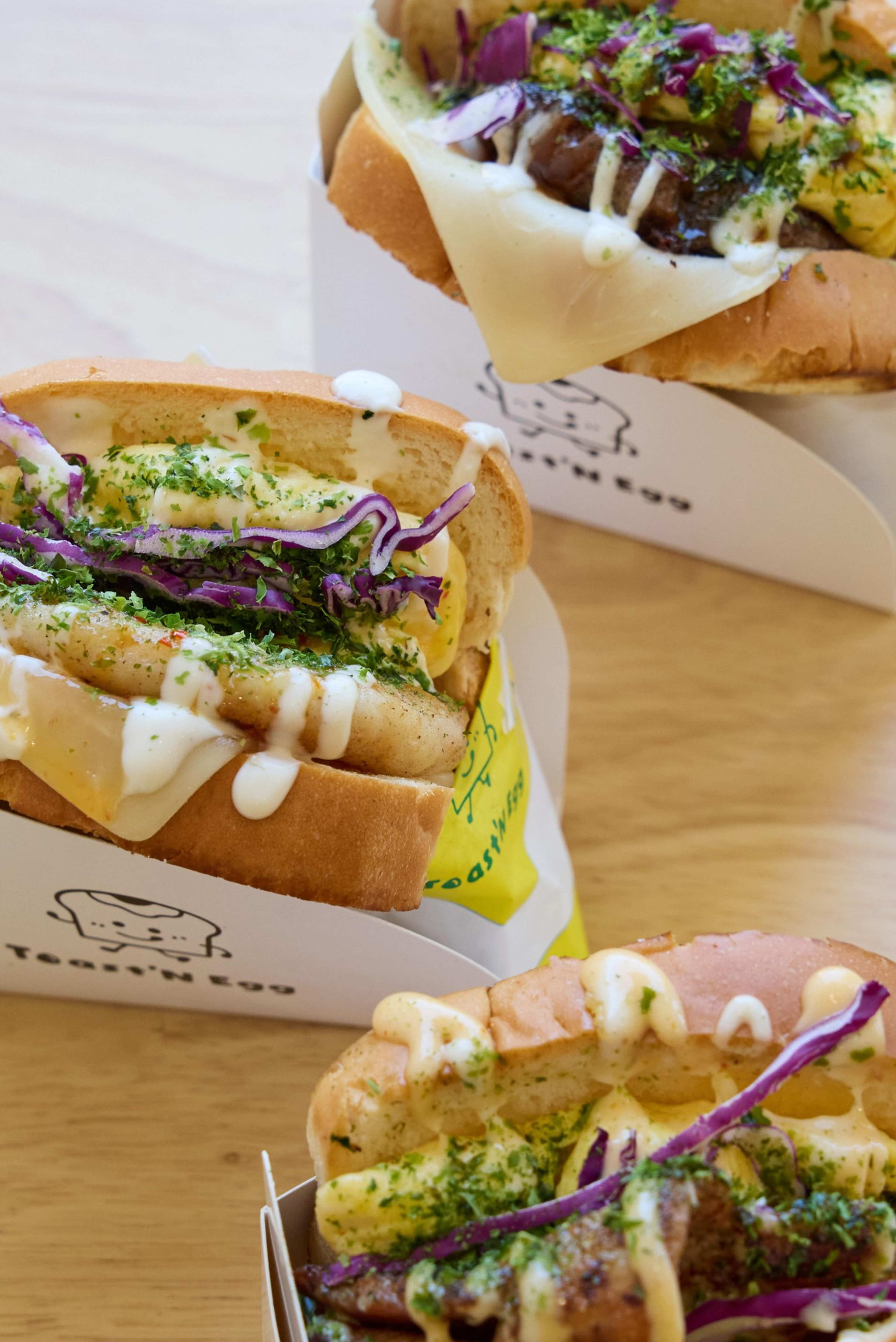 Three egg sandwiches are shown with fluffy bread, cooked eggs, cheese, shredded purple cabbage, and creamy sauces. They are placed in white paper holders displaying a cartoon toast.