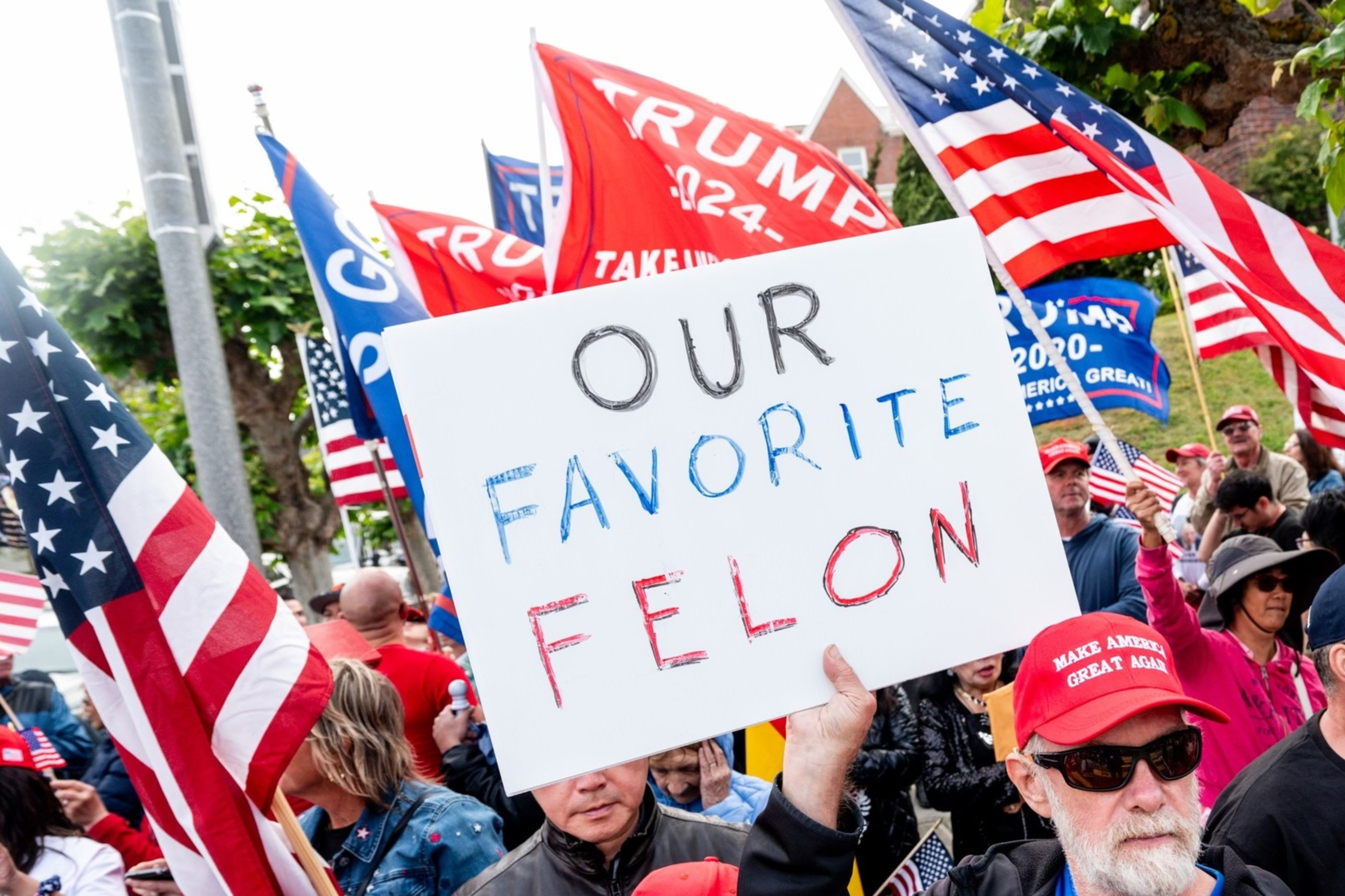A crowd of people, many wearing red "Make America Great Again" hats, hold American flags and a sign that reads "OUR FAVORITE FELON" at a rally.