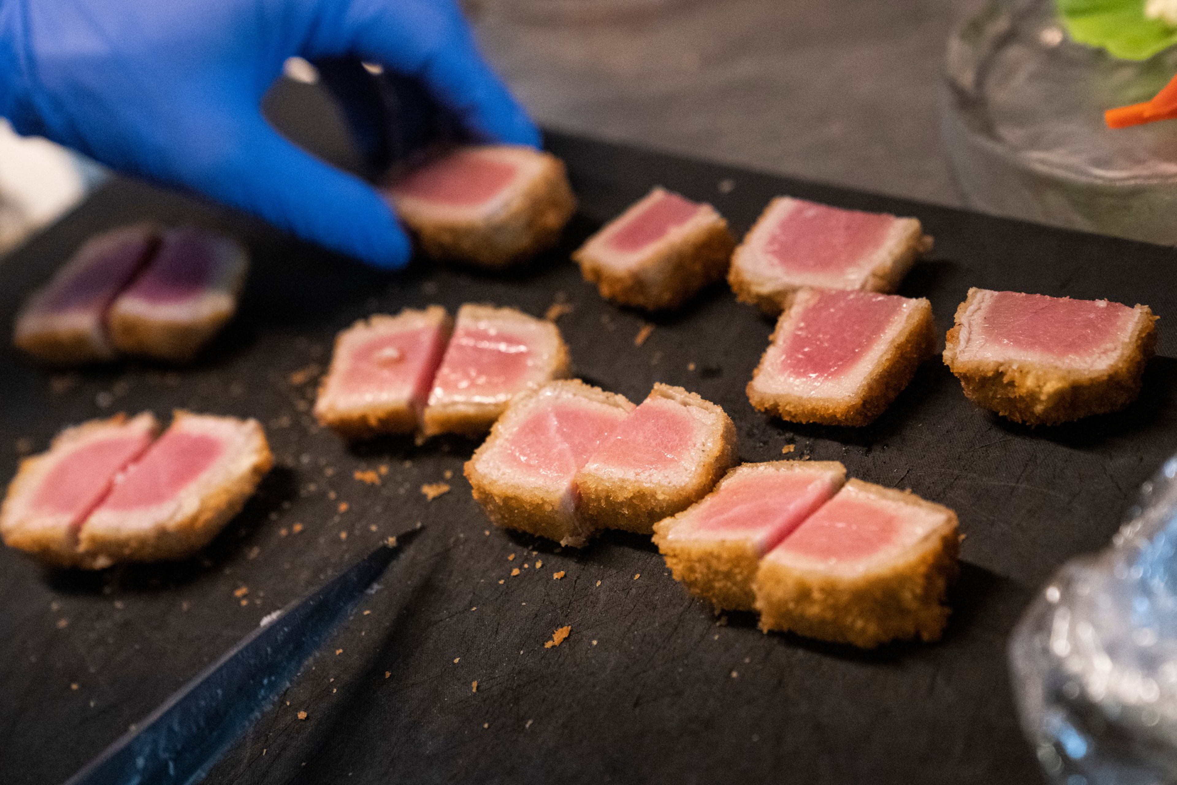 A gloved hand arranges breaded and partially seared tuna pieces on a black cutting board. The tuna is pink inside and golden brown outside.