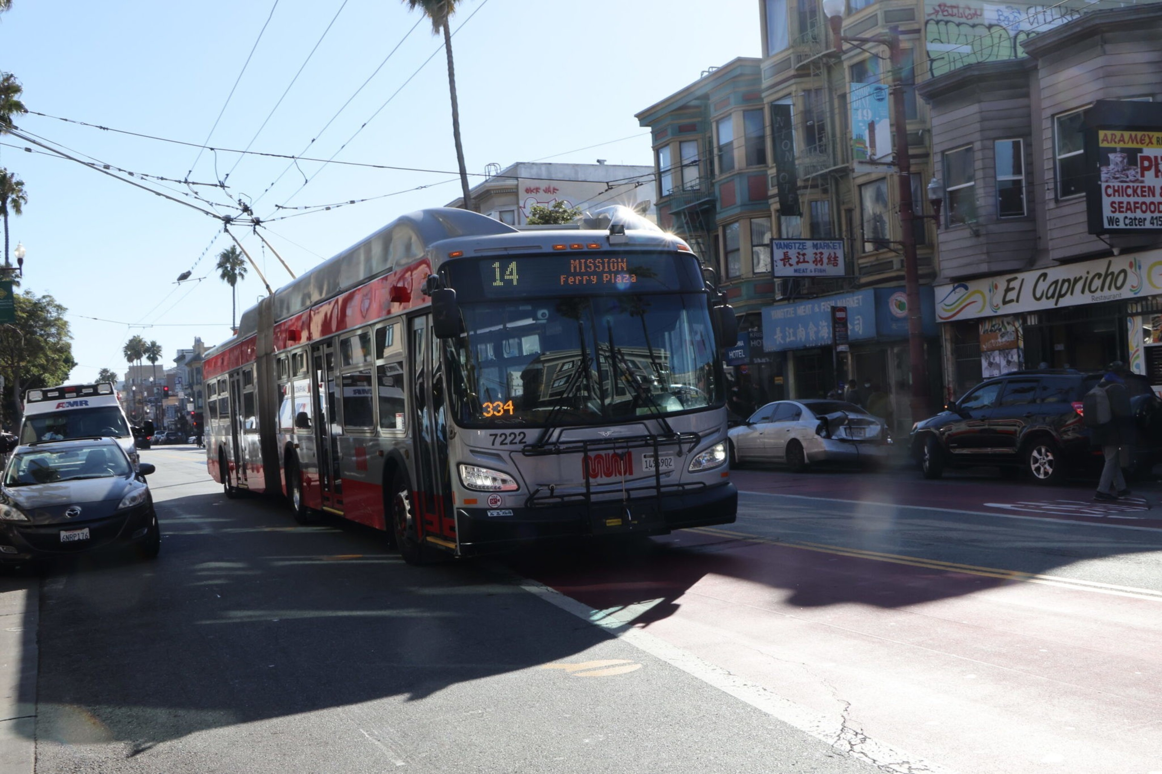 A red and silver articulated bus marked "14 Mission Ferry Plaza" drives along a street lined with parked cars and various storefronts.
